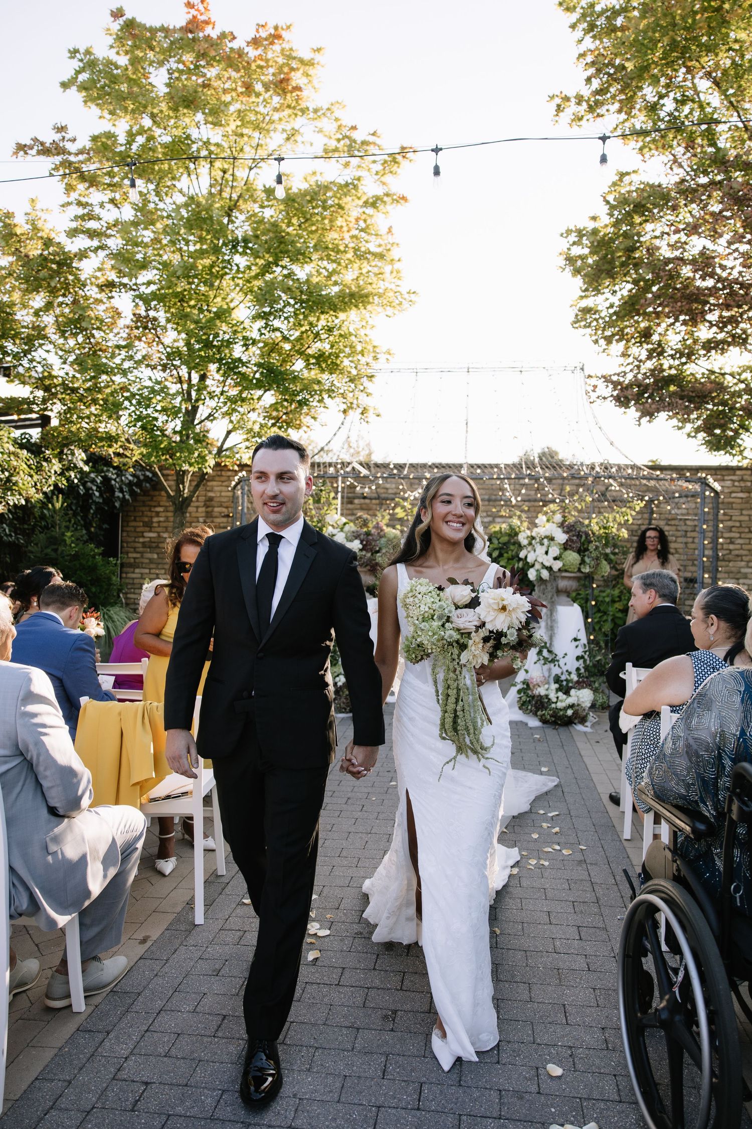 Bride and groom walk down outdoor wedding aisle, bride holding white floral bouquet, guests seated on both sides.