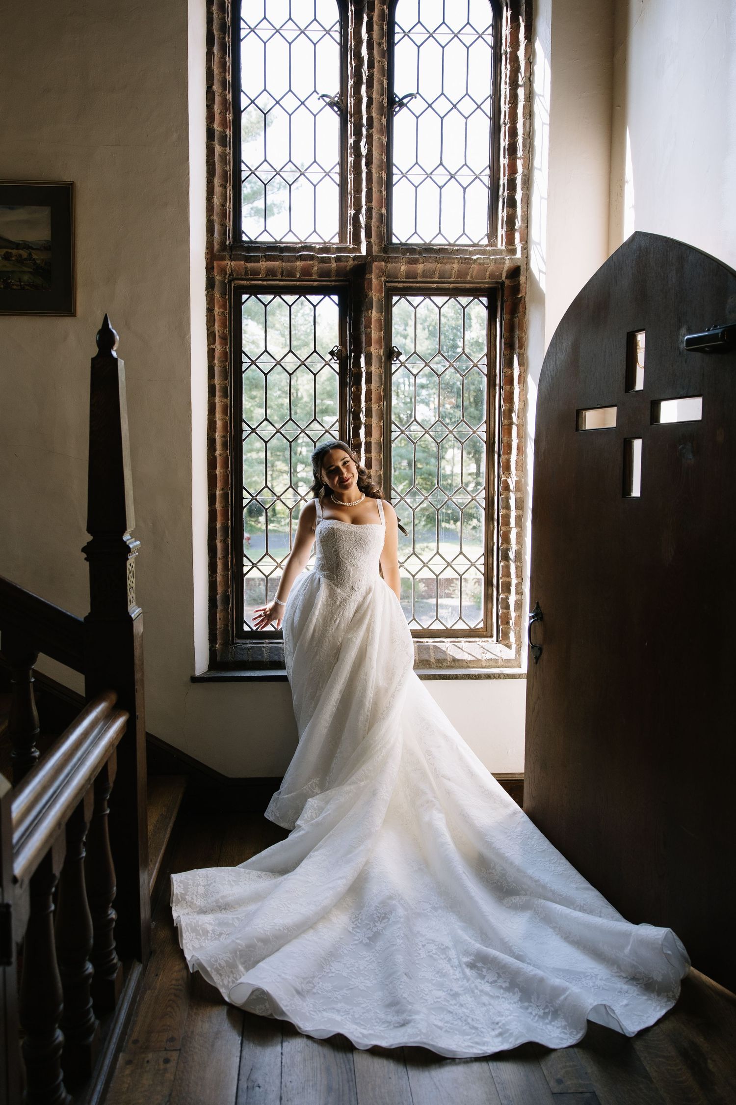 Bride in elegant white wedding gown with long train stands by ornate leaded glass windows in historic venue.
