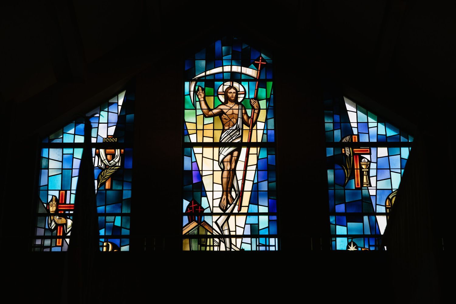Colorful stained glass church window depicting the resurrection of Jesus Christ flanked by saints in blue tones.