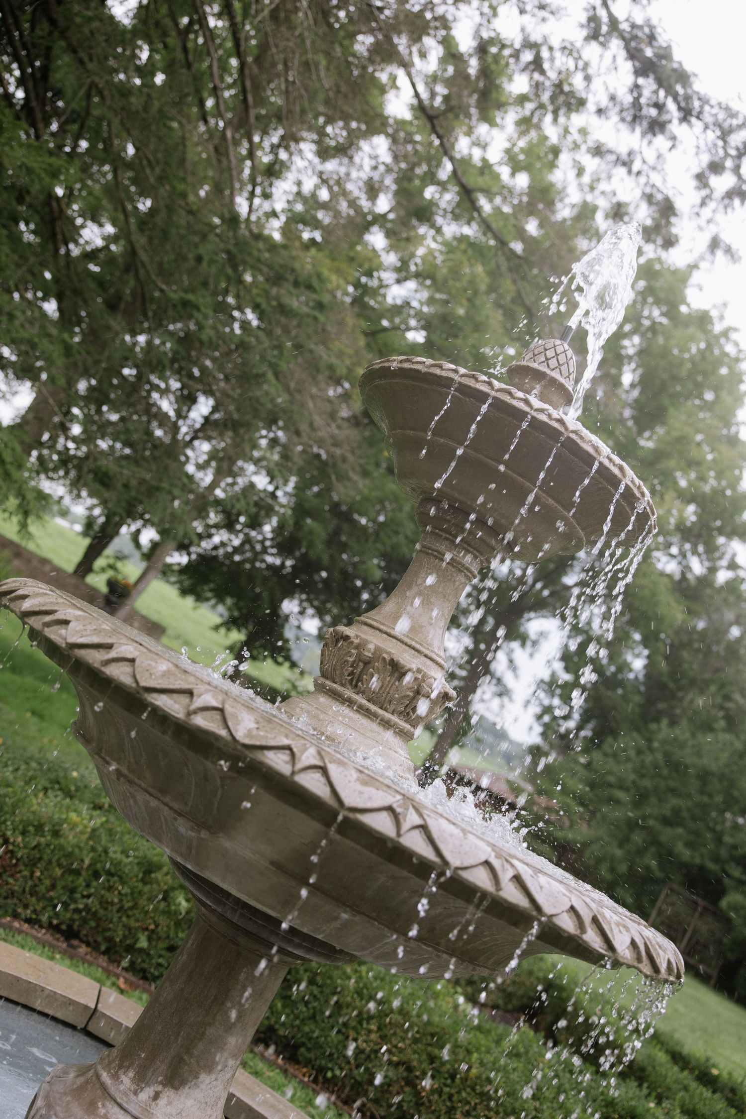 A multi-tiered stone garden fountain with cascading water streams, surrounded by lush green trees and hedges.