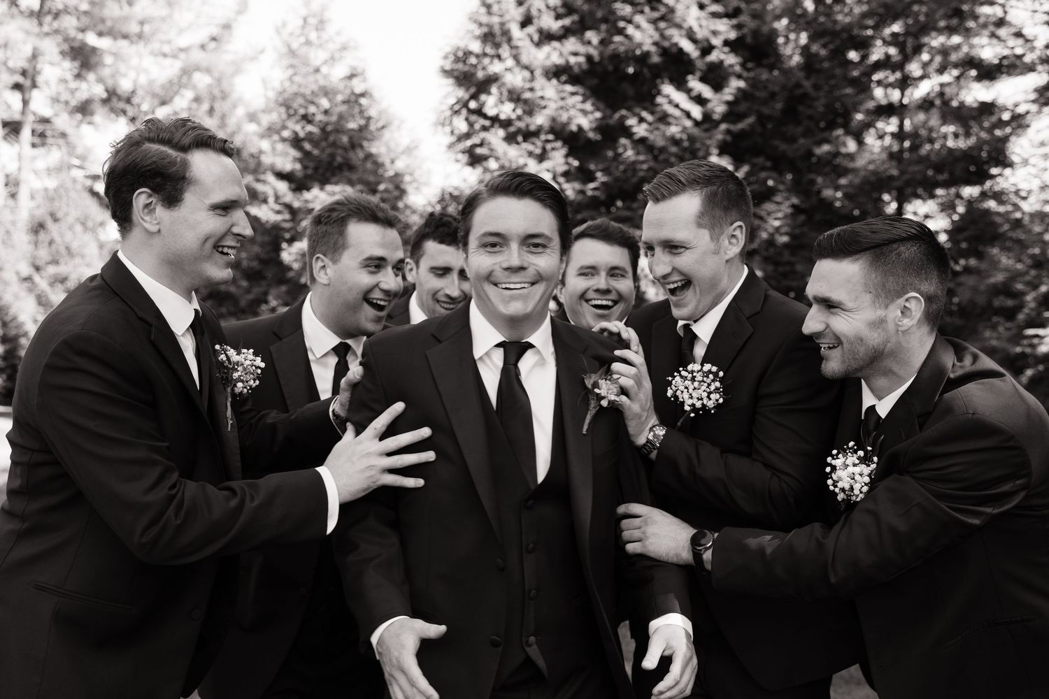 Black and white photo of a groom surrounded by laughing groomsmen in suits with boutonnieres at a wedding.
