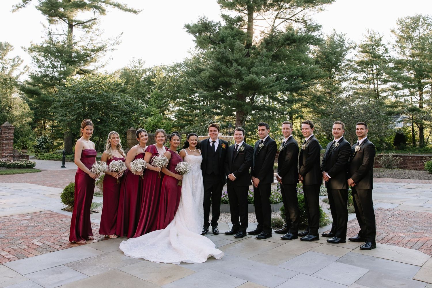 Wedding party poses outdoors, bridesmaids in burgundy gowns and groomsmen in black suits flanking bride and groom.