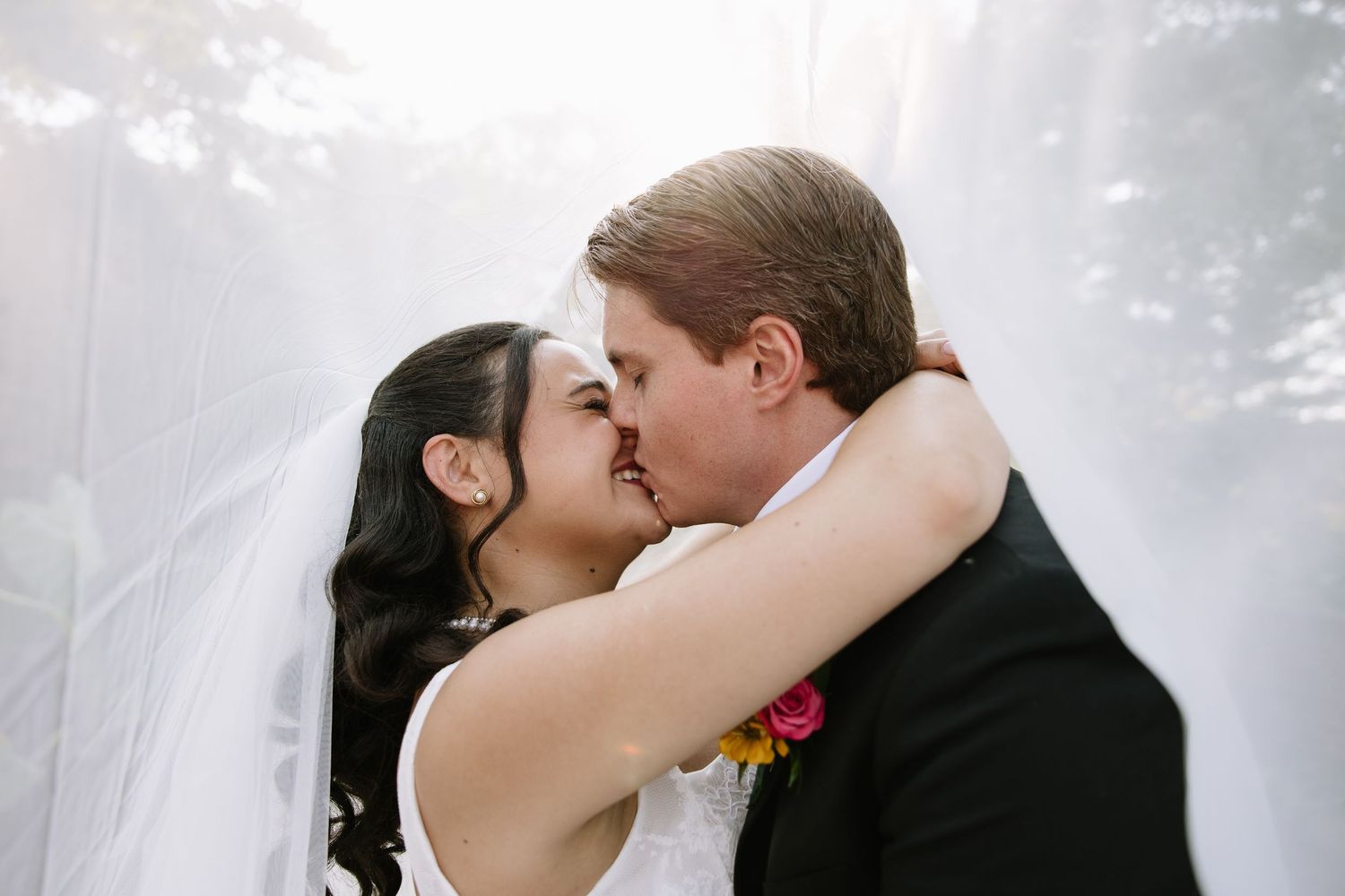 Bride and groom sharing a romantic kiss under a flowing white veil on their wedding day.