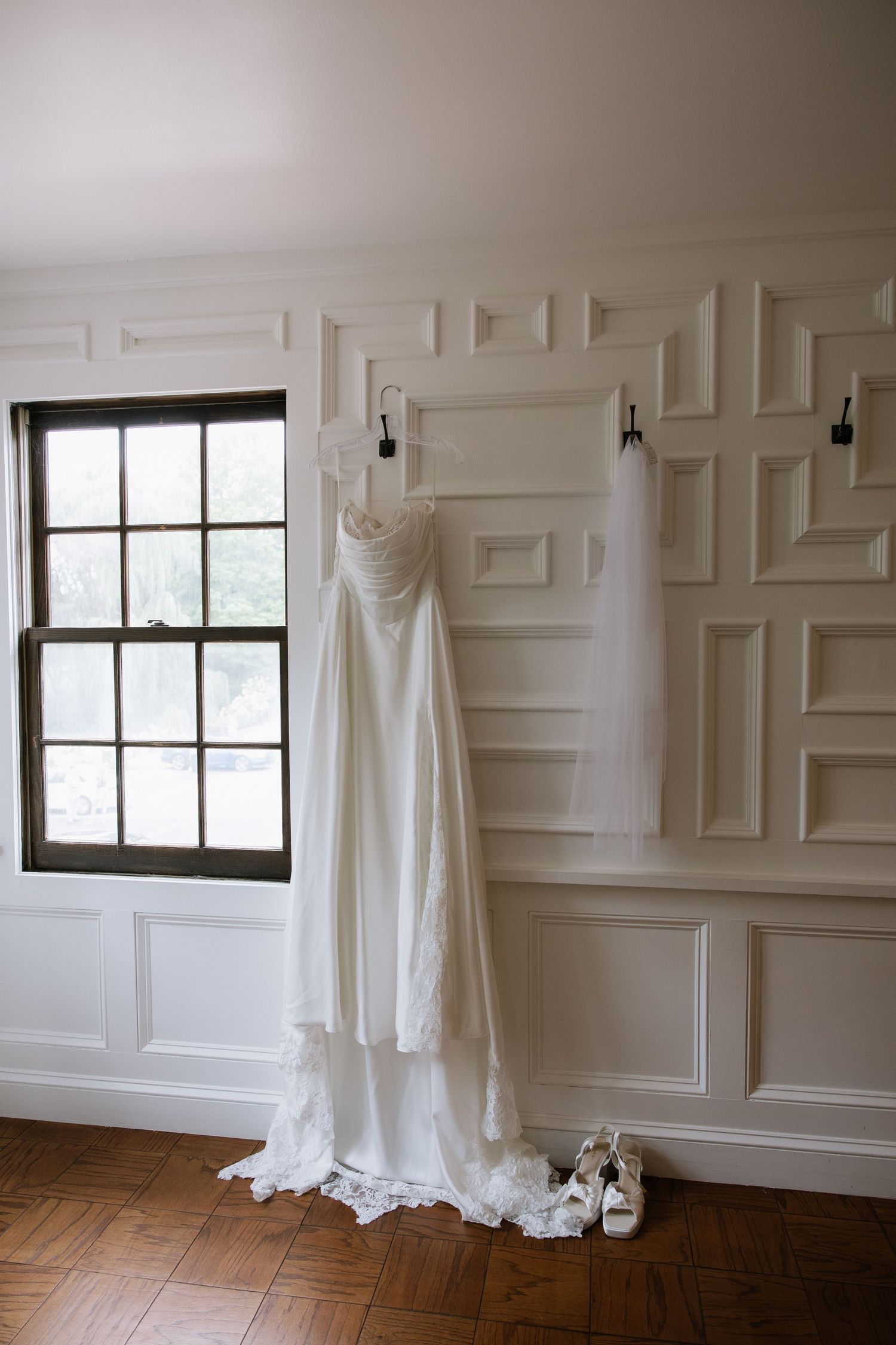 White wedding dress and veil hanging on wall hooks beside a window, with bridal heels on hardwood floor.
