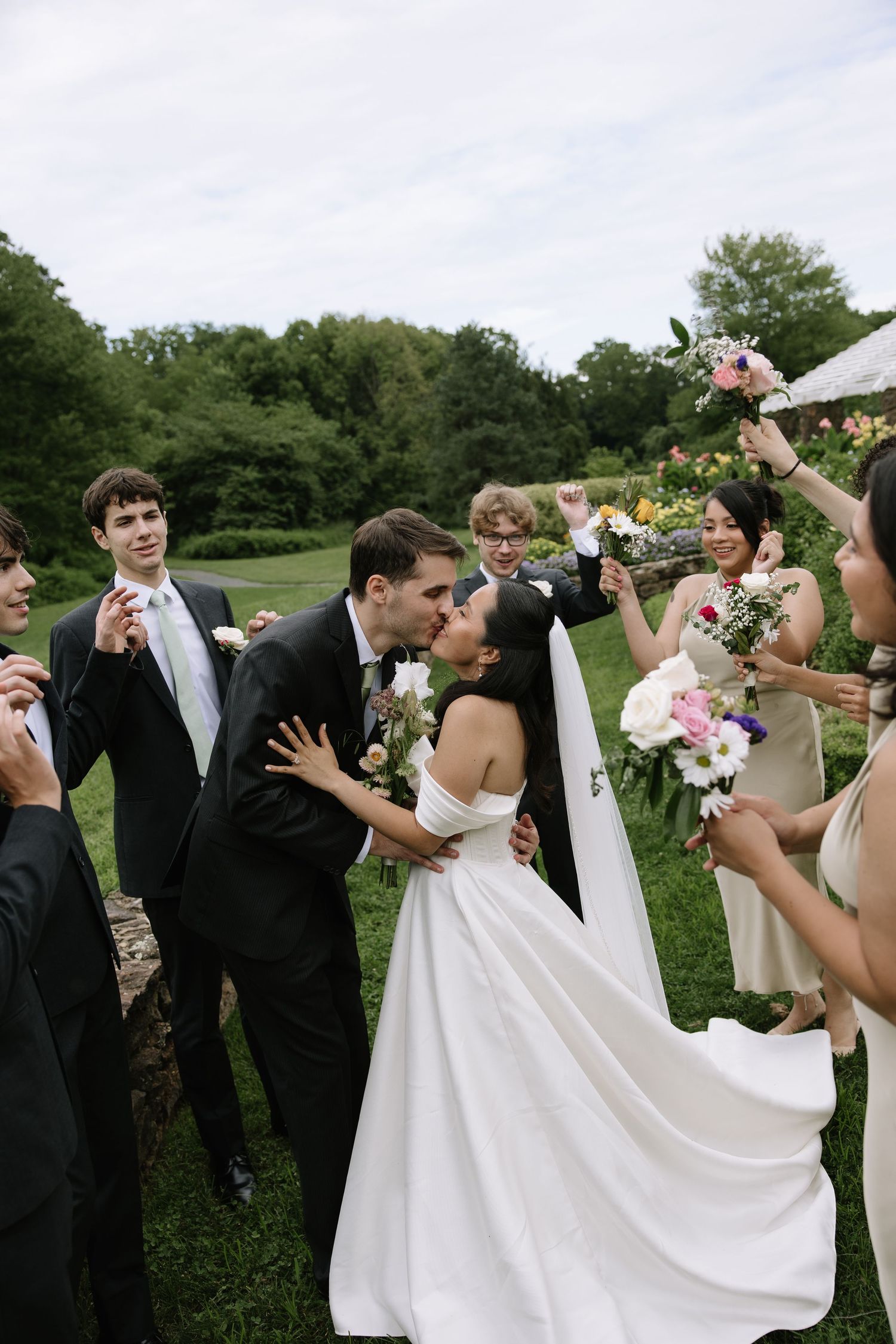 A bride and groom share a kiss surrounded by their wedding party holding bouquets outdoors on a lush green lawn.