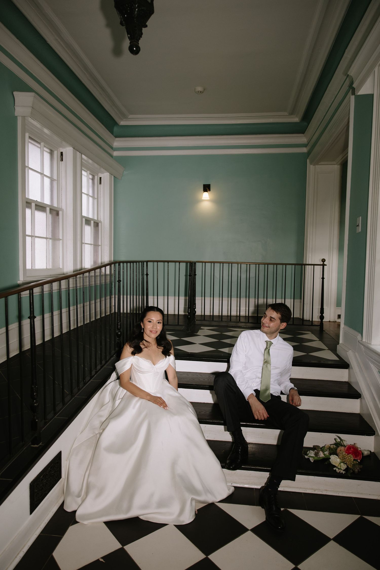 Bride and groom sitting on black and white checkered stairs in elegant teal hallway during wedding portrait session.