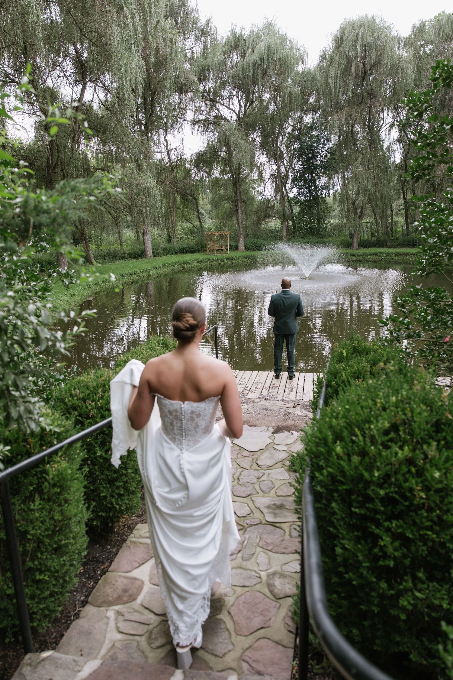 Bride in white gown walks toward groom by a serene pond with fountain, surrounded by lush willow trees.