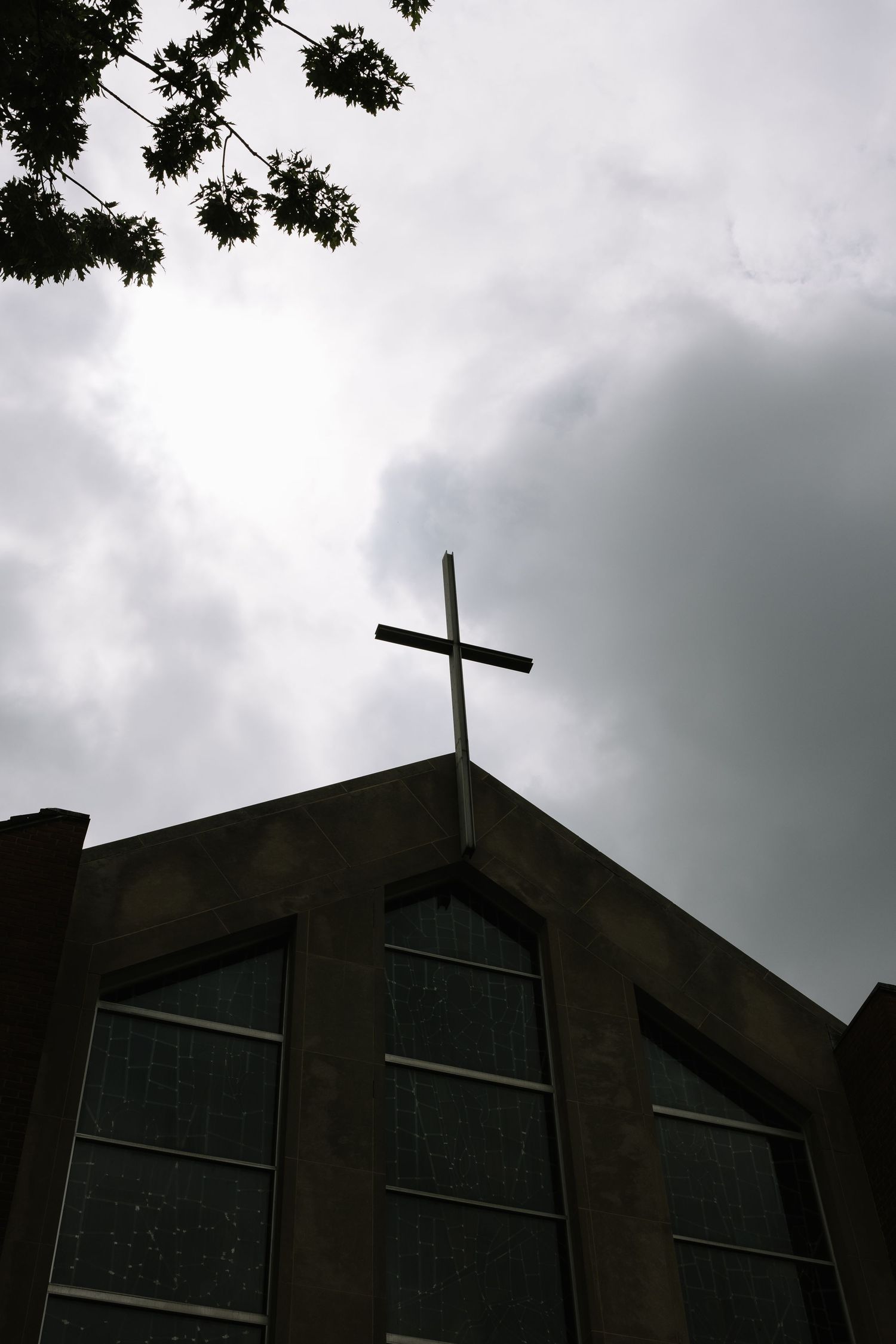 A Christian church steeple with a cross silhouetted against a dramatic cloudy sky, framed by tree branches.