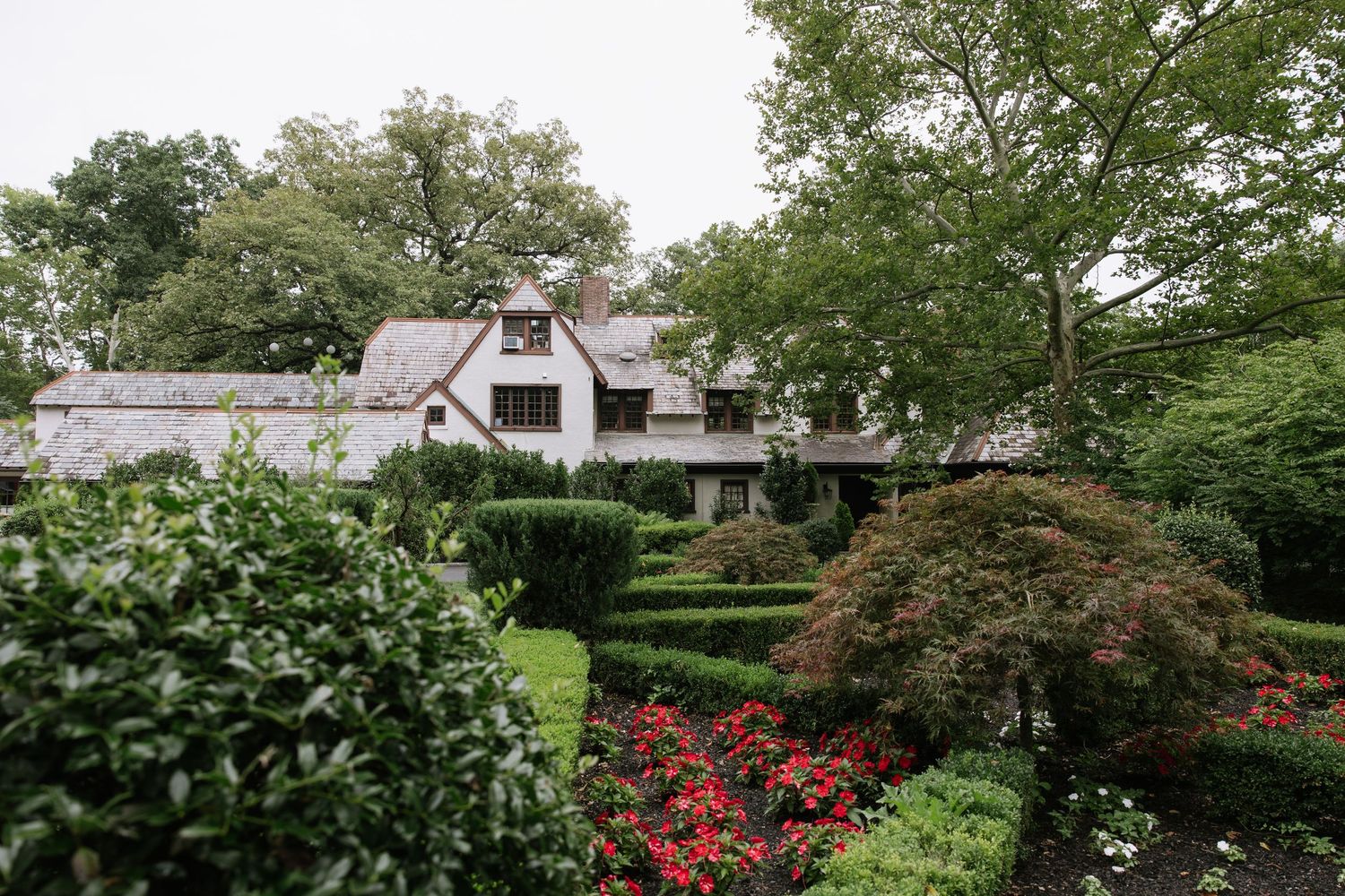 Tudor-style home surrounded by lush manicured hedges, red flowering shrubs, and tall green trees in an elegant garden setting.
