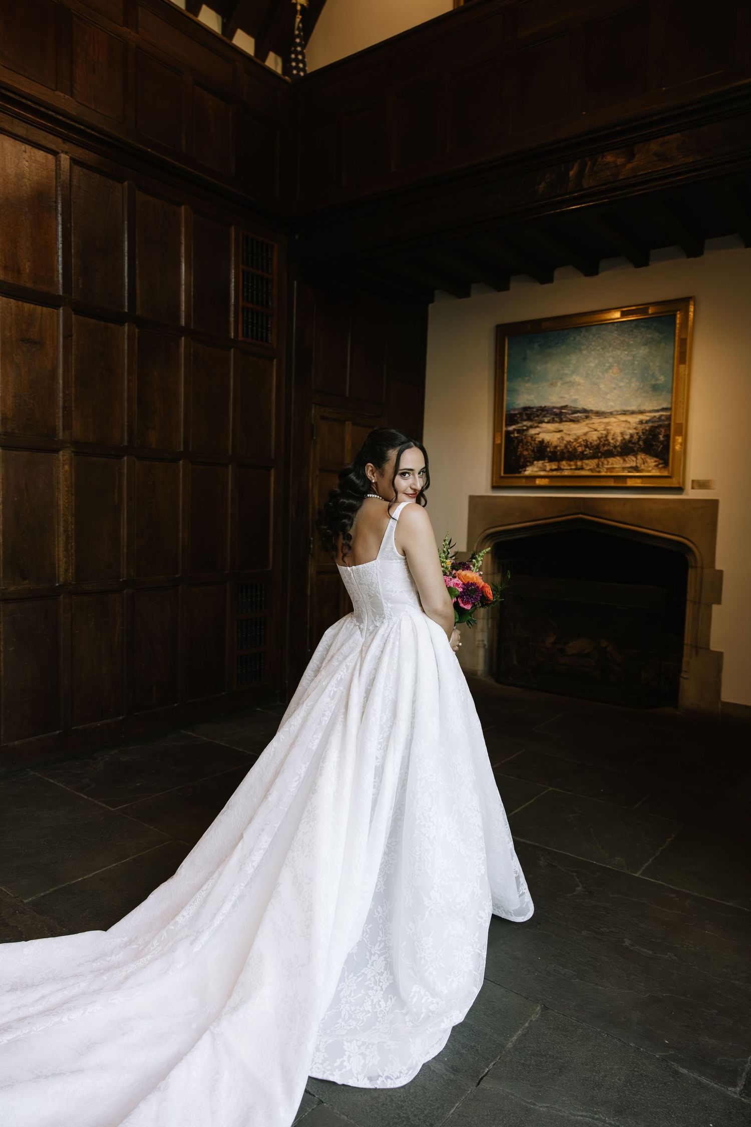 Bride in elegant white lace gown with long train holds bouquet, posing near fireplace in wood-paneled room.