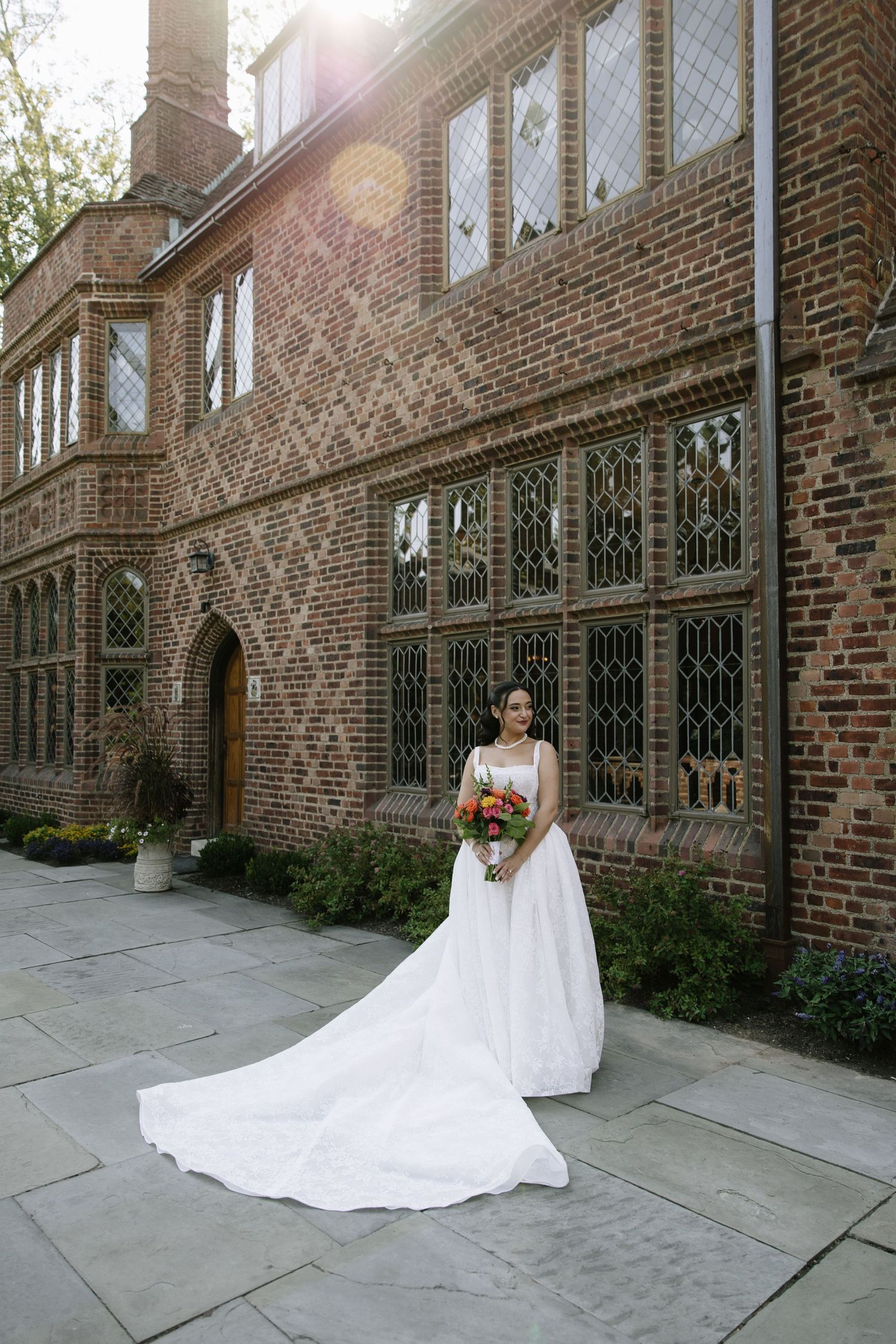 Bride in white ball gown with long train holds bouquet outside elegant brick manor with leaded glass windows.