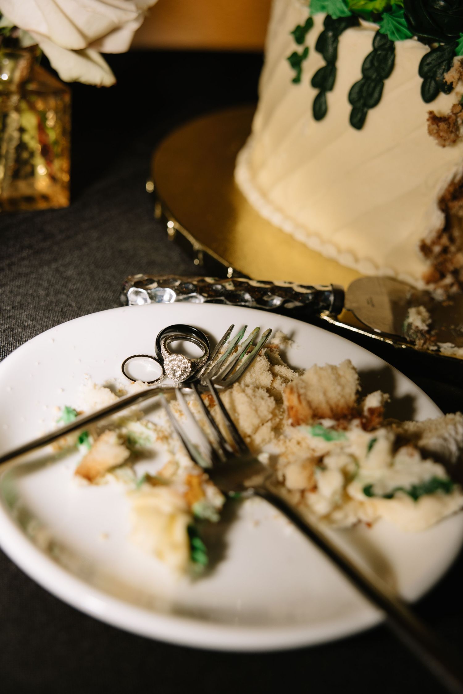 Wedding rings resting on plate of eaten cake slices beside decorated wedding cake.