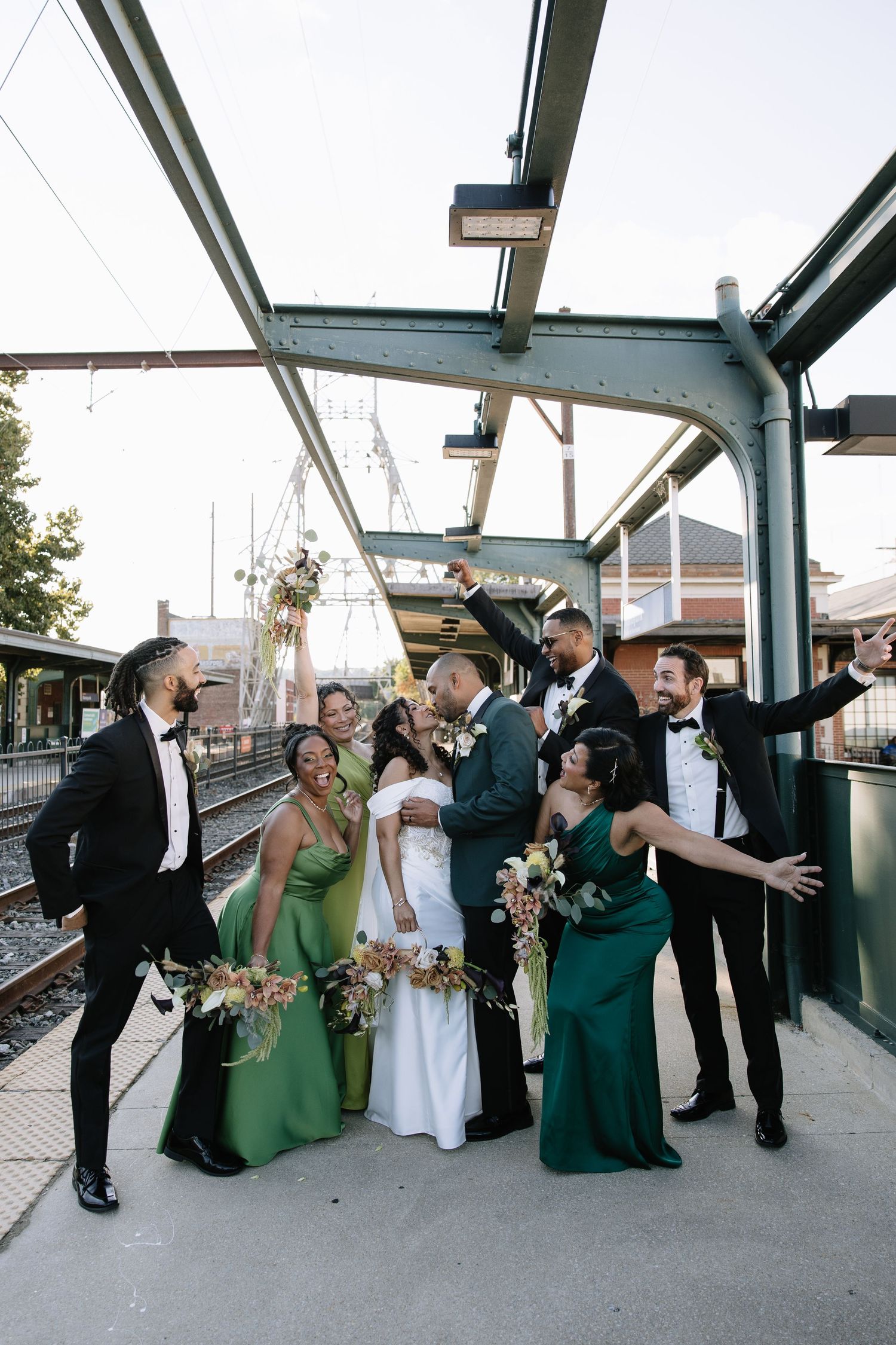 Wedding party celebrates on train platform, bridesmaids in green gowns and groomsmen in tuxedos cheering with bride and groom.