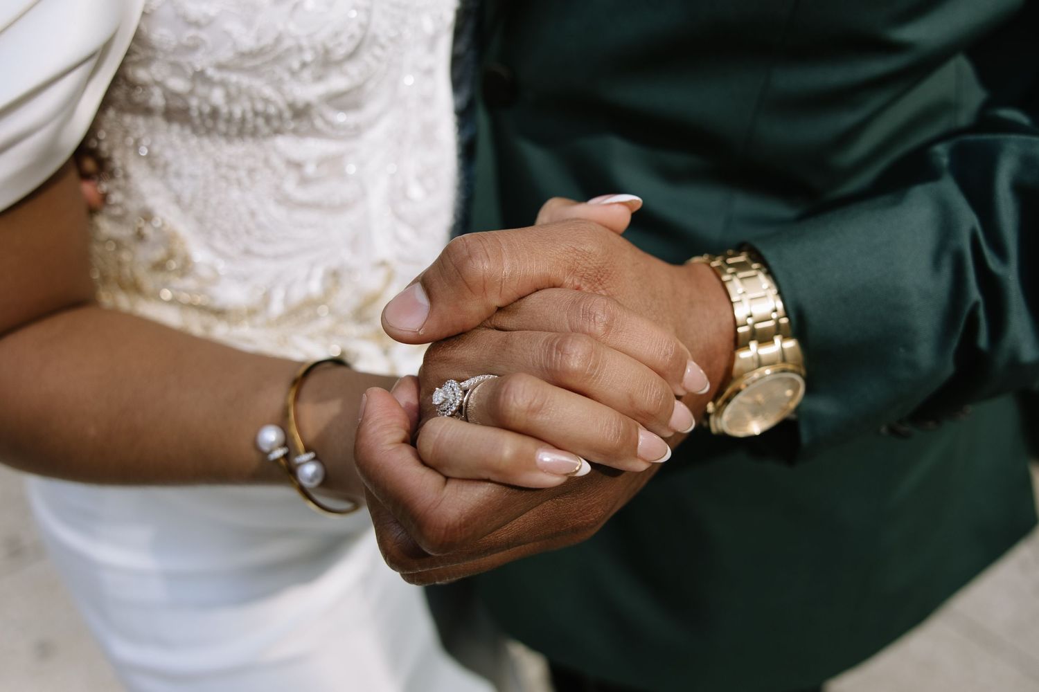 Close-up of newlyweds holding hands, bride wearing diamond ring and bracelet, groom in green suit with gold watch.