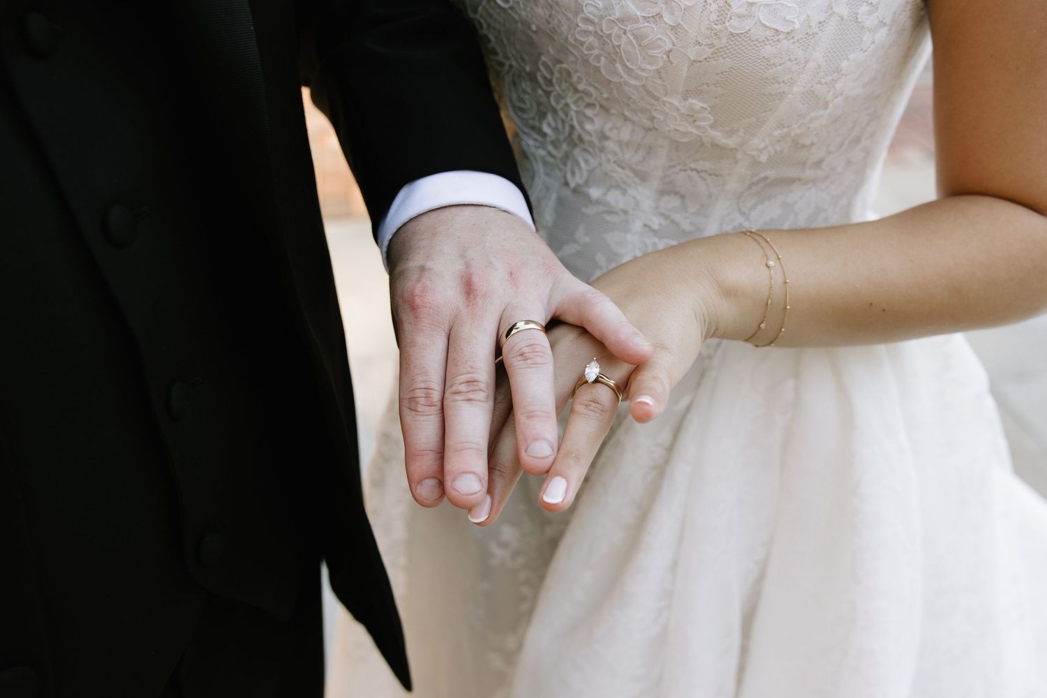 Newlywed couple holding hands showing wedding rings, bride in lace dress and groom in black suit.