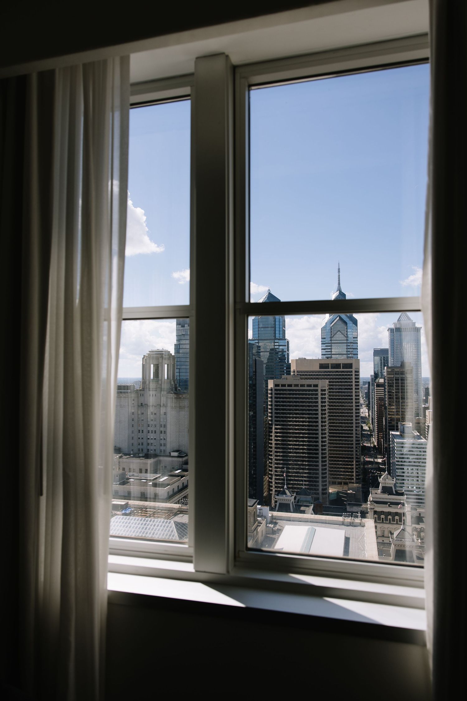 City skyline view through a hotel window with white curtains, featuring modern skyscrapers under a blue sky.
