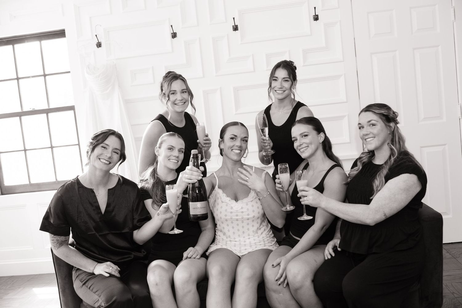 Black and white photo of a bride in white dress celebrating with seven bridesmaids in black, toasting with champagne glasses.