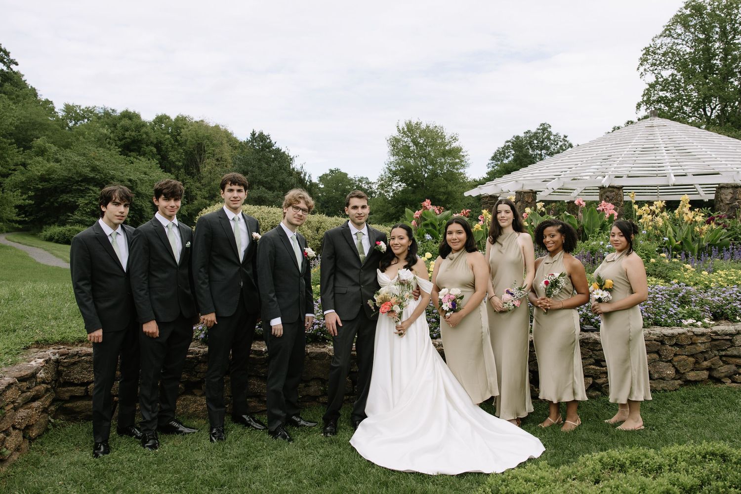 Wedding party poses outdoors, groomsmen in black suits and bridesmaids in champagne dresses flanking bride and groom.