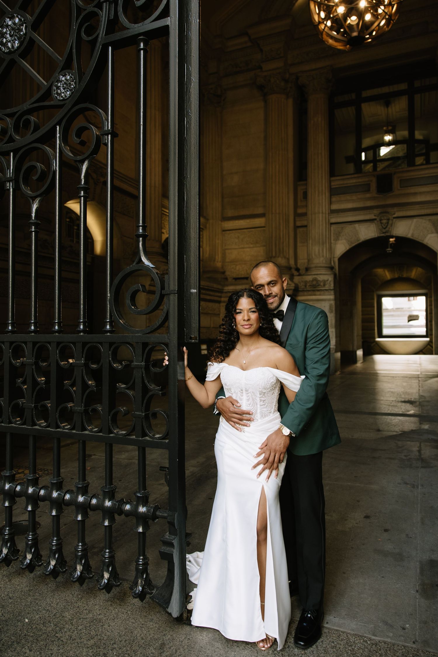 Elegant couple posing by ornate iron gates, bride in white off-shoulder gown, groom in green tuxedo jacket.