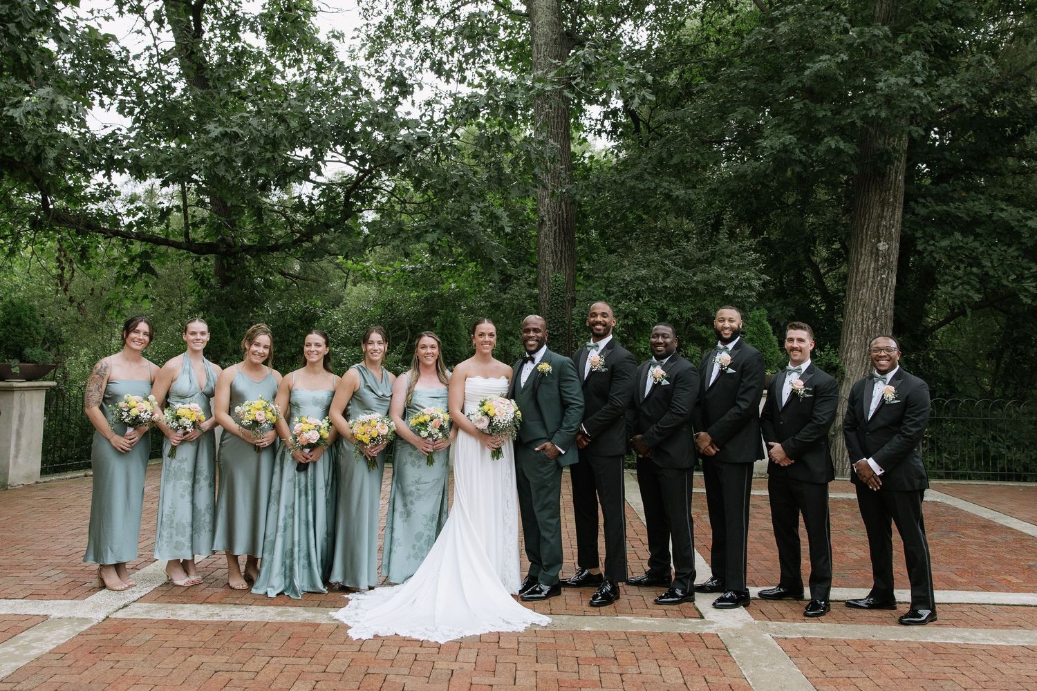 Wedding party poses outdoors, bridesmaids in sage green gowns with yellow bouquets, groomsmen in black suits, bride and groom centered.