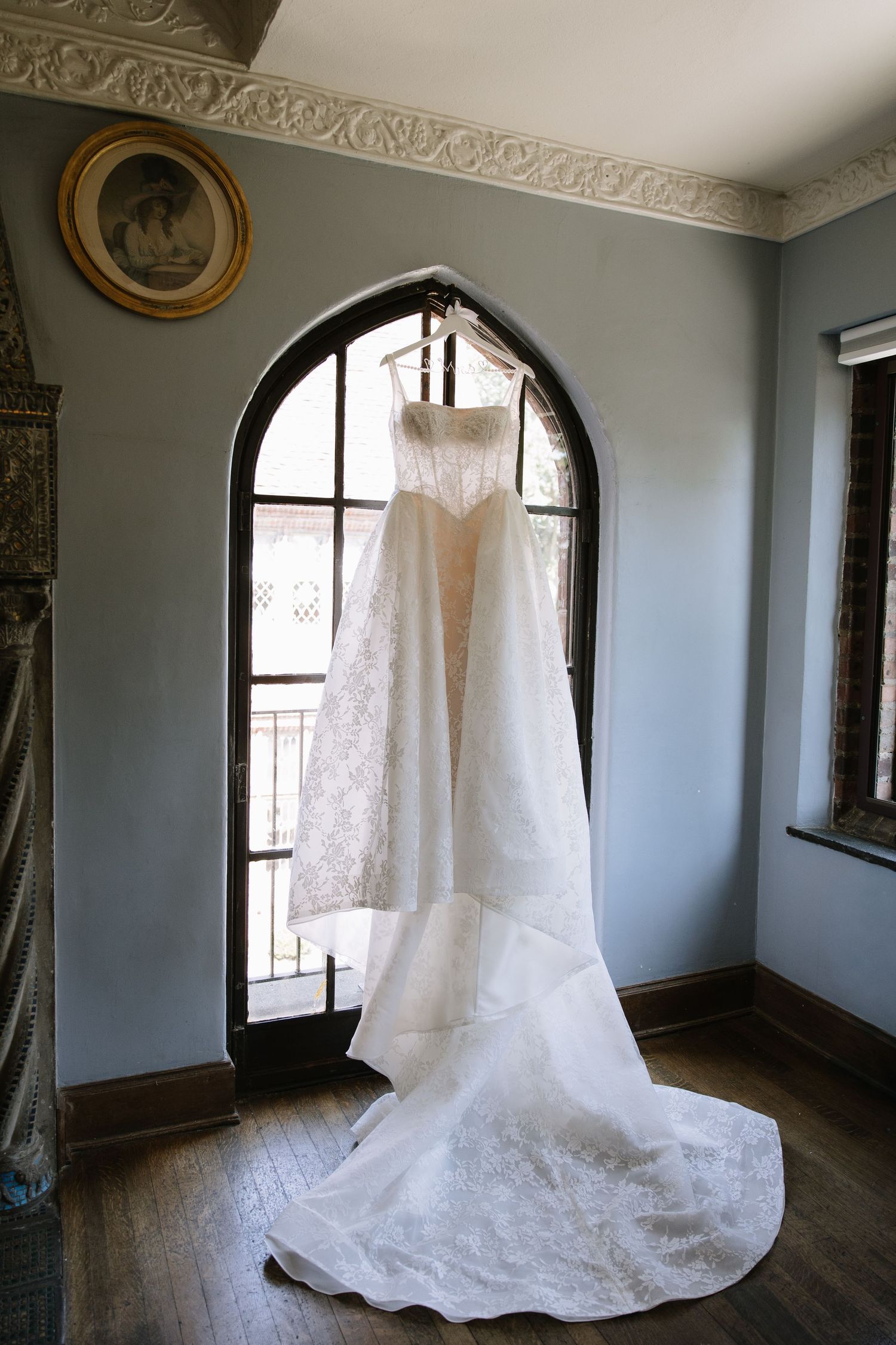 Elegant white lace wedding dress with a long train hanging in an arched window of a vintage blue room.