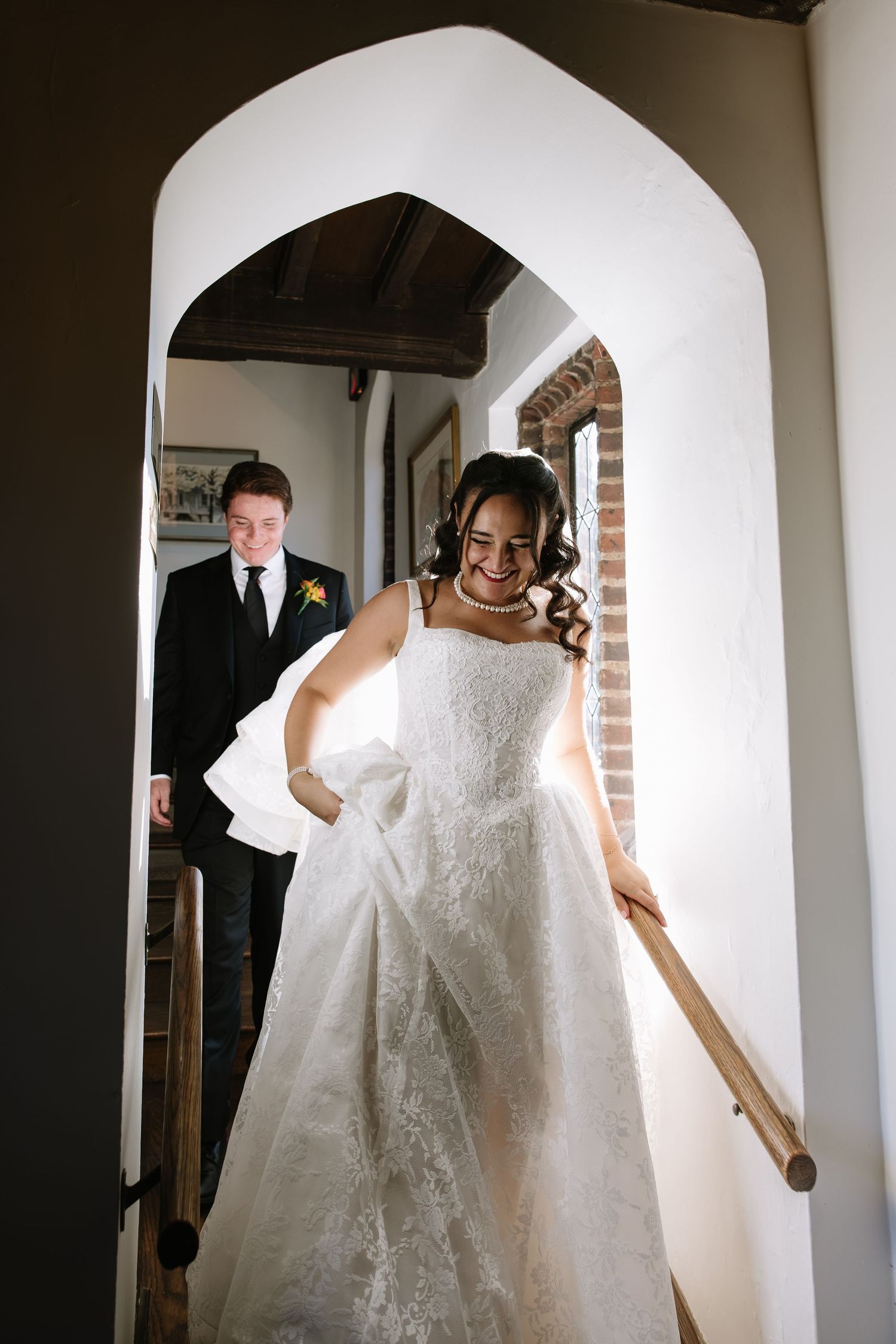 Smiling bride in lace ball gown descends staircase through arched doorway, groom in black suit follows behind.