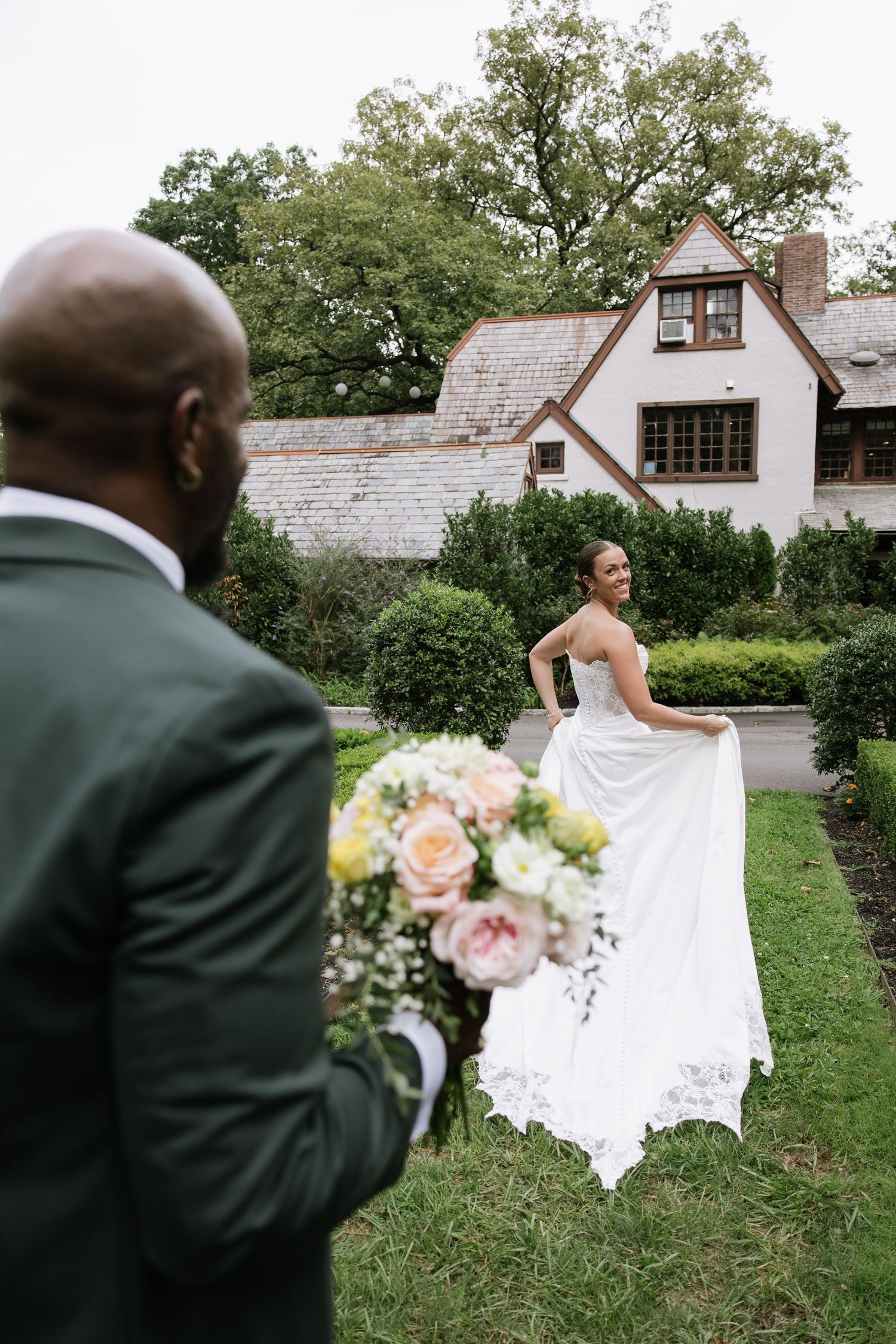 Groom in green suit holds bouquet as bride in white gown smiles in garden near Tudor-style home.