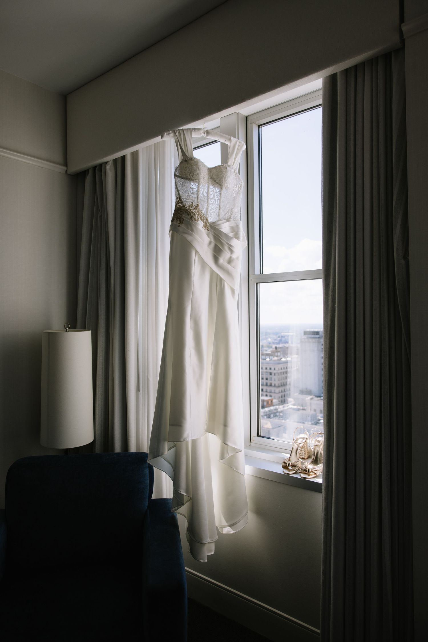 White wedding dress hanging in hotel window with city skyline view.