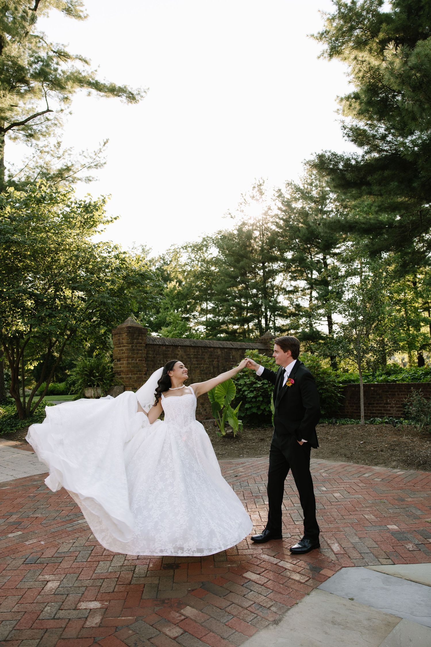 Bride in white gown twirls as groom in black tuxedo holds her hand on brick patio surrounded by lush green trees.