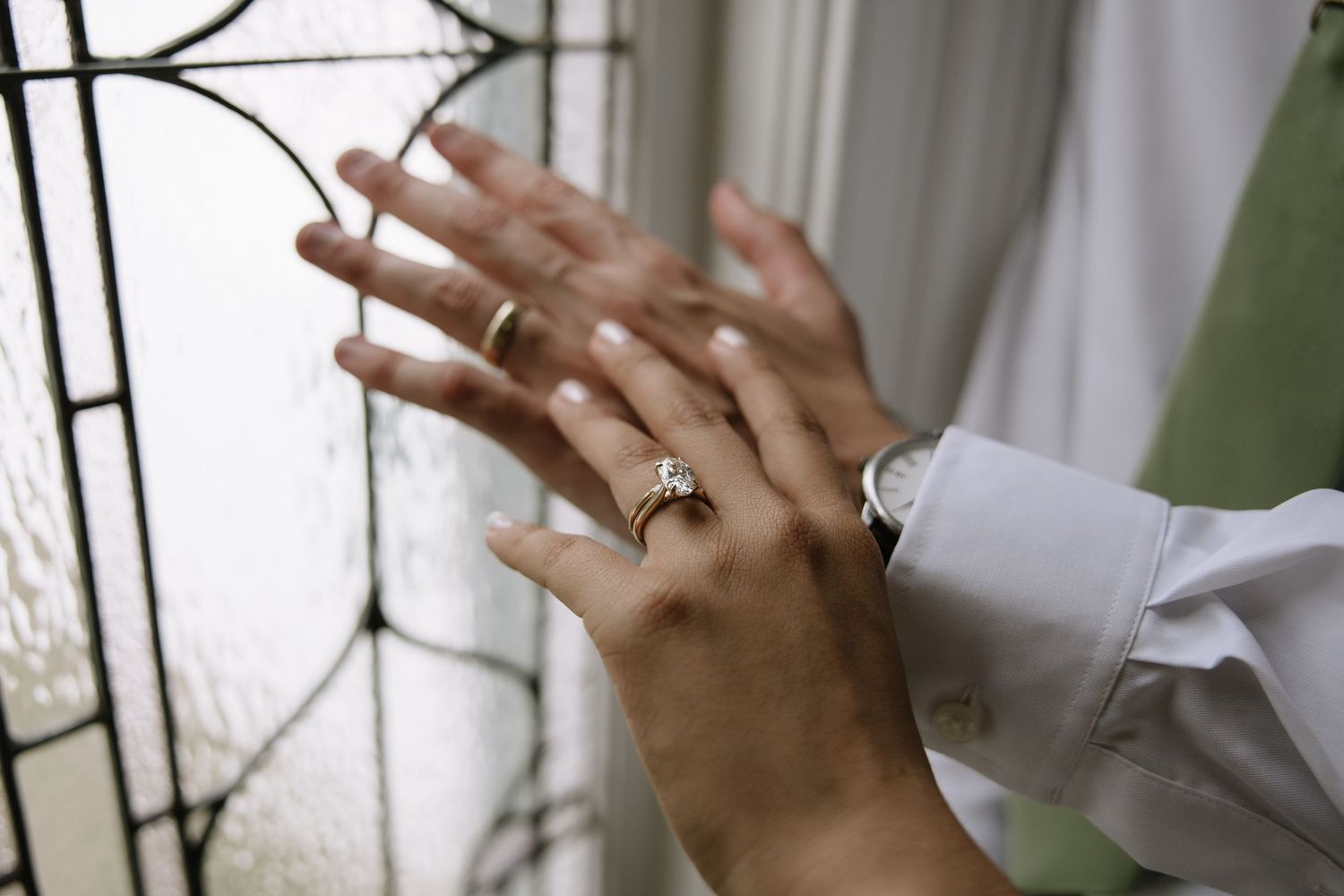 Close-up of hands near stained glass window, one wearing a diamond engagement ring and wedding band.