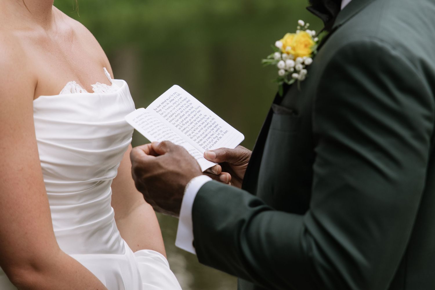 Groom in green suit reads wedding vows from paper to bride in white strapless gown during outdoor ceremony.