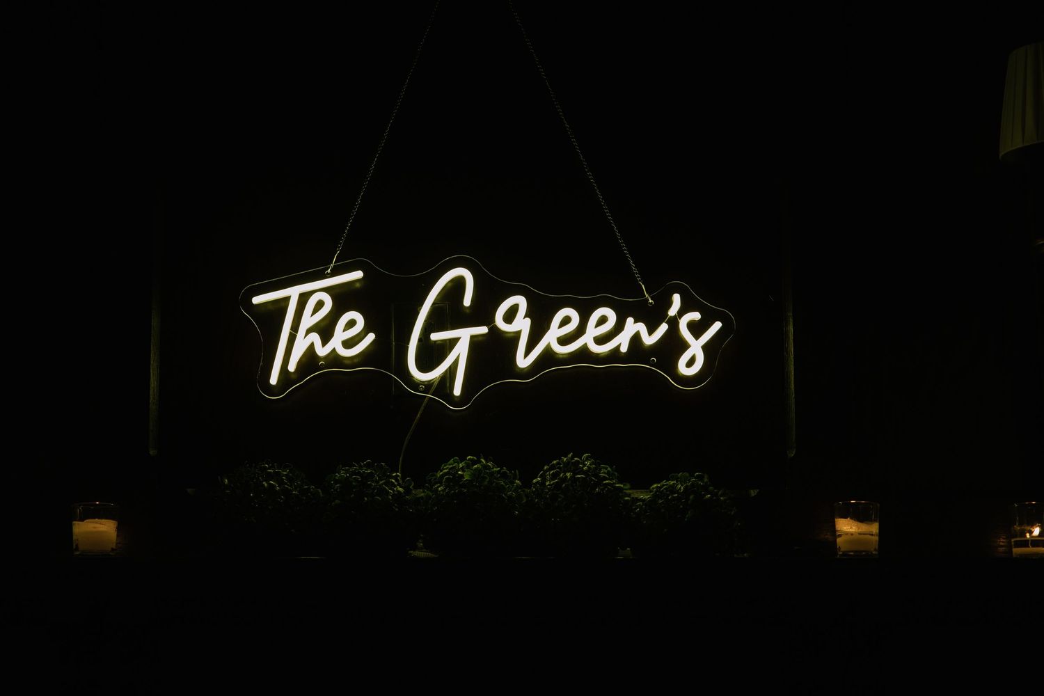 Glowing neon sign reading The Green's in warm white cursive script, hanging in darkness above lush greenery.