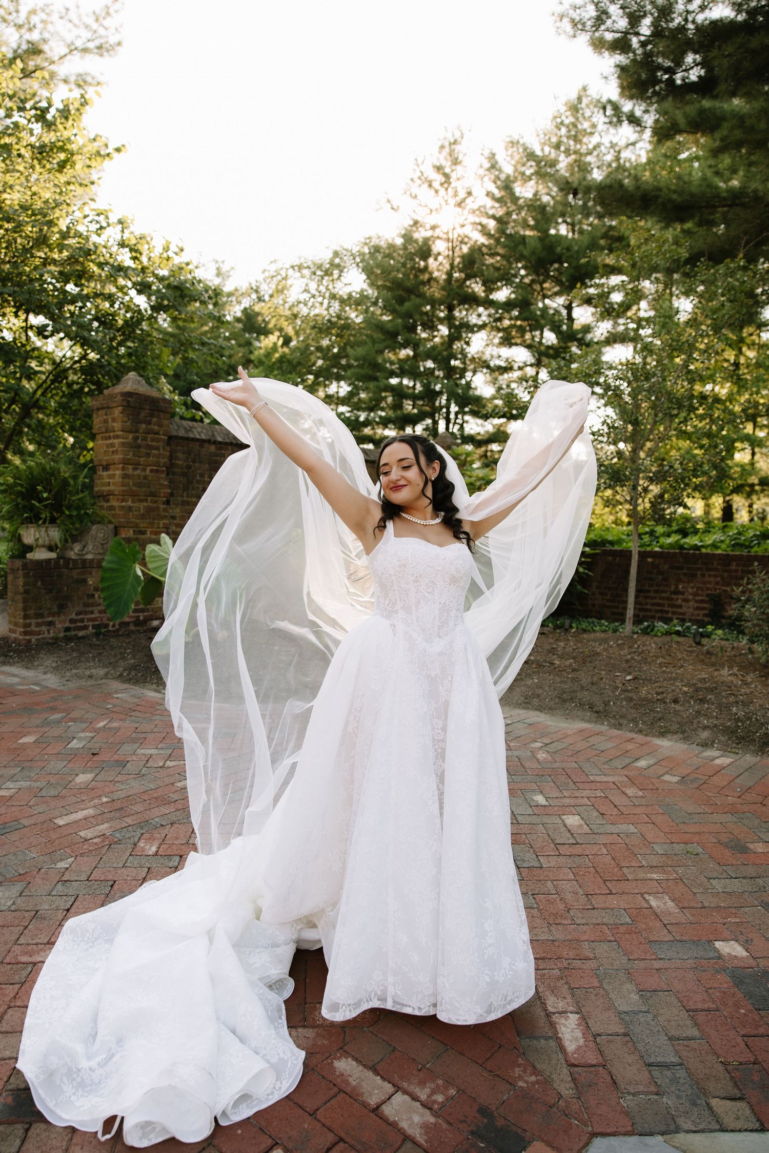 Bride in white wedding gown twirling veil outdoors on brick patio surrounded by lush green trees.