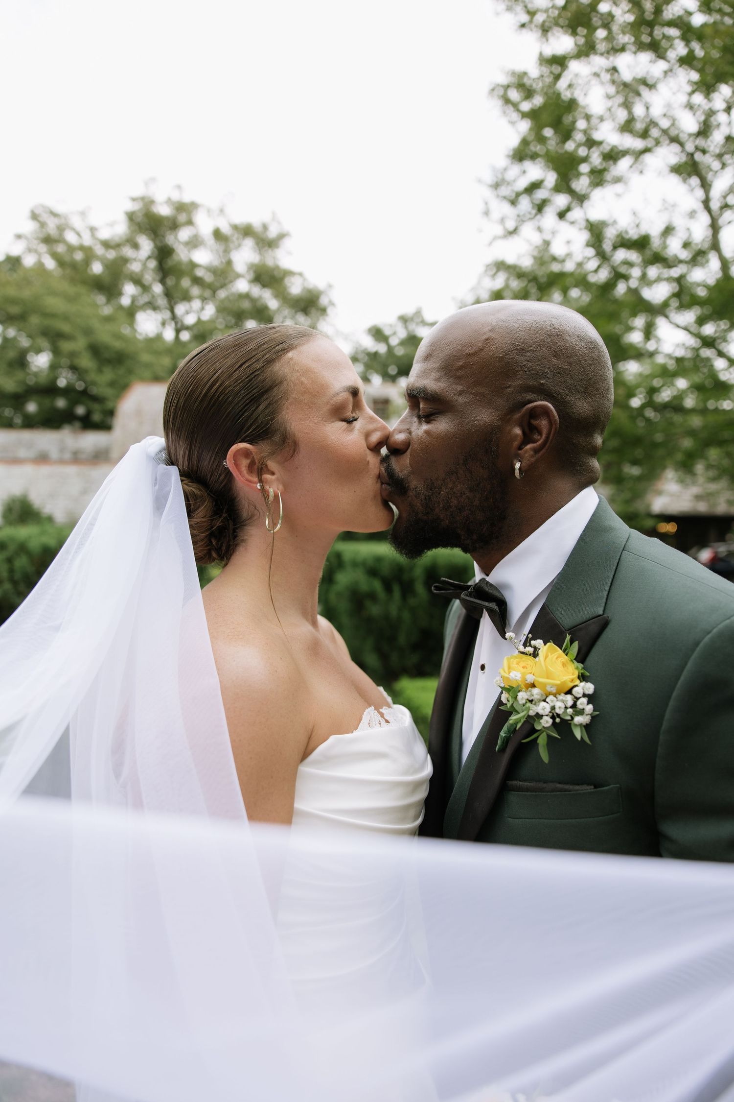 Bride and groom sharing a kiss outdoors, groom in green tuxedo with yellow boutonniere, bride in white gown with veil.