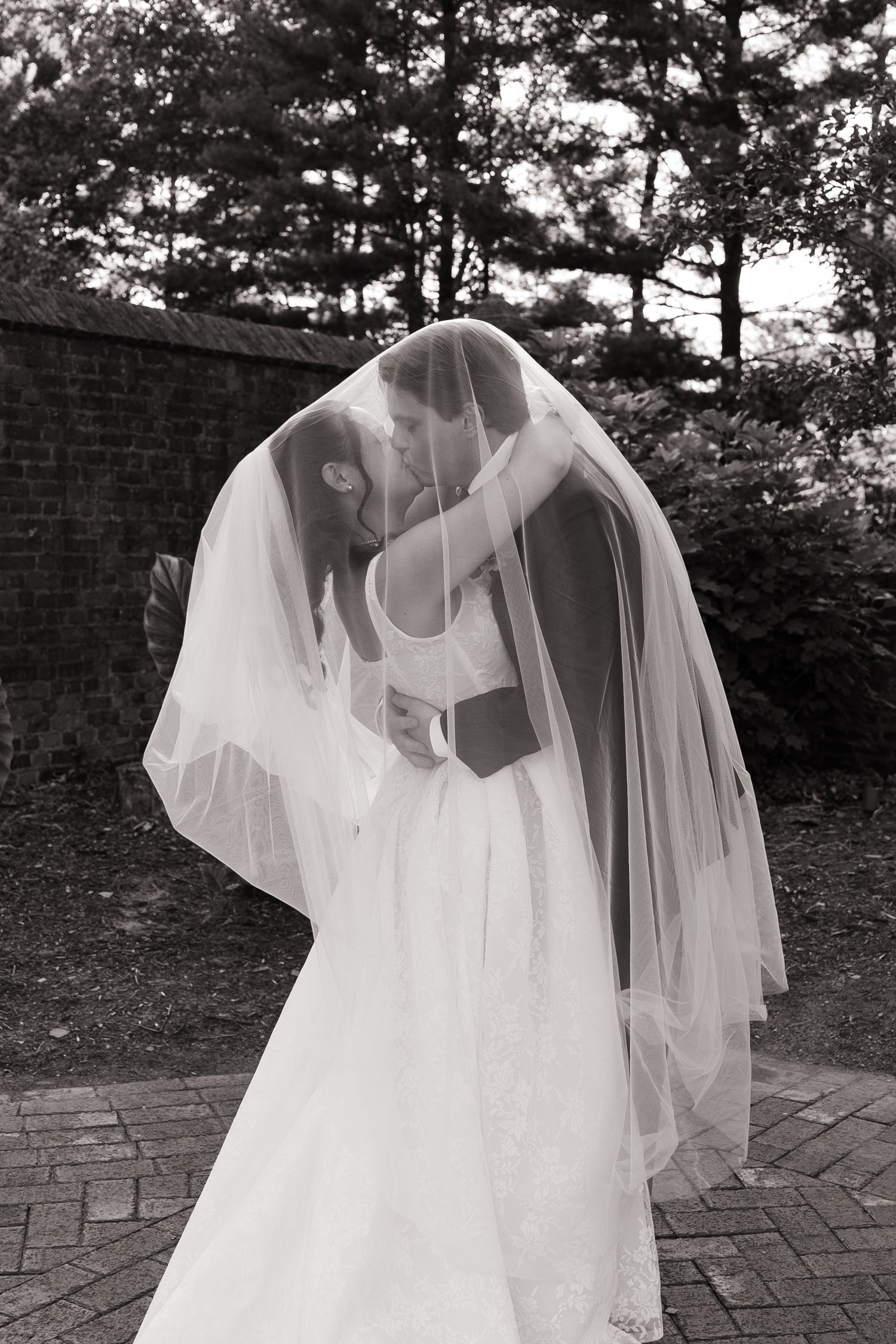Black and white photo of bride and groom sharing a kiss under a wedding veil outdoors.