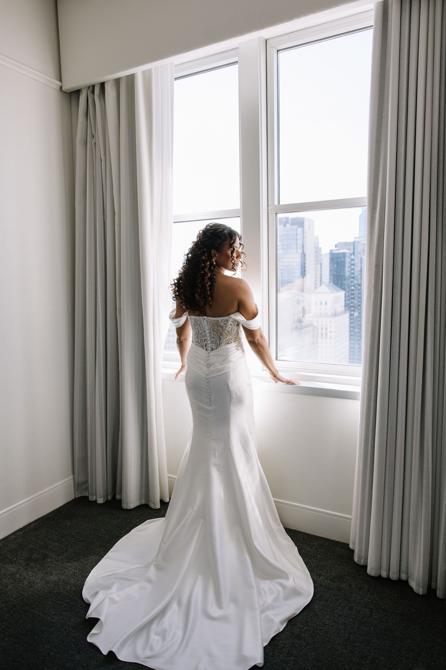 Bride in white off-shoulder mermaid gown with lace corset back stands at window overlooking city skyline.