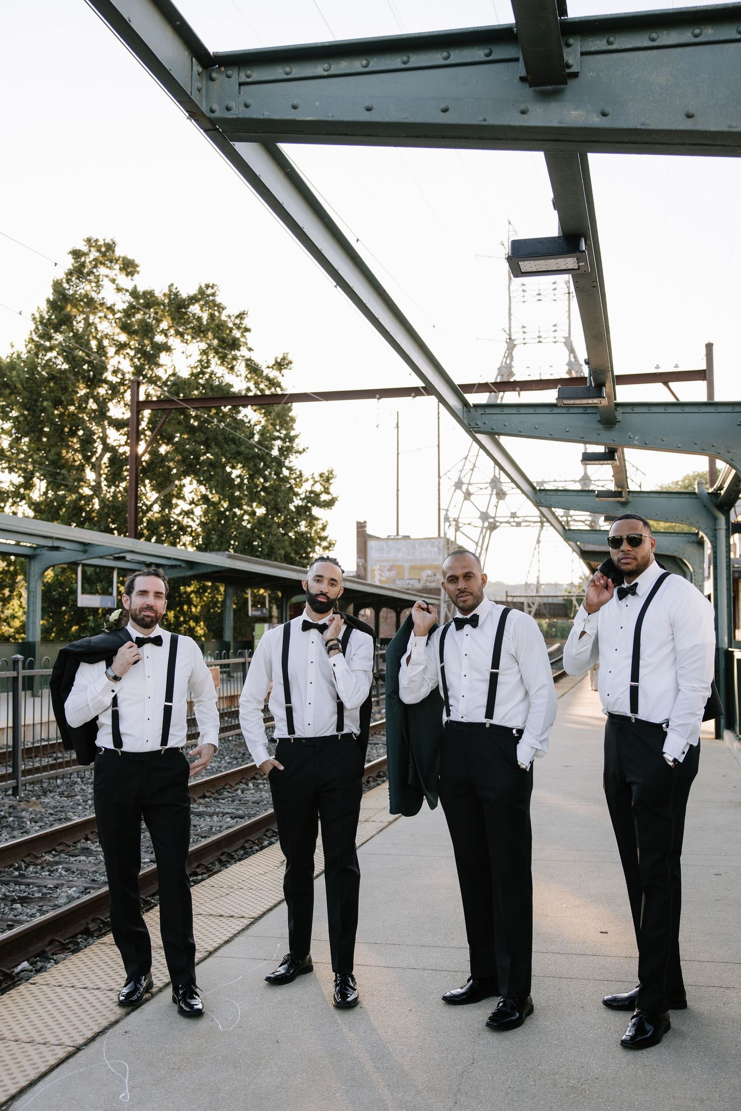 Groomsmen in white shirts, black bow ties, and suspenders pose on a train platform for a stylish wedding party photo.