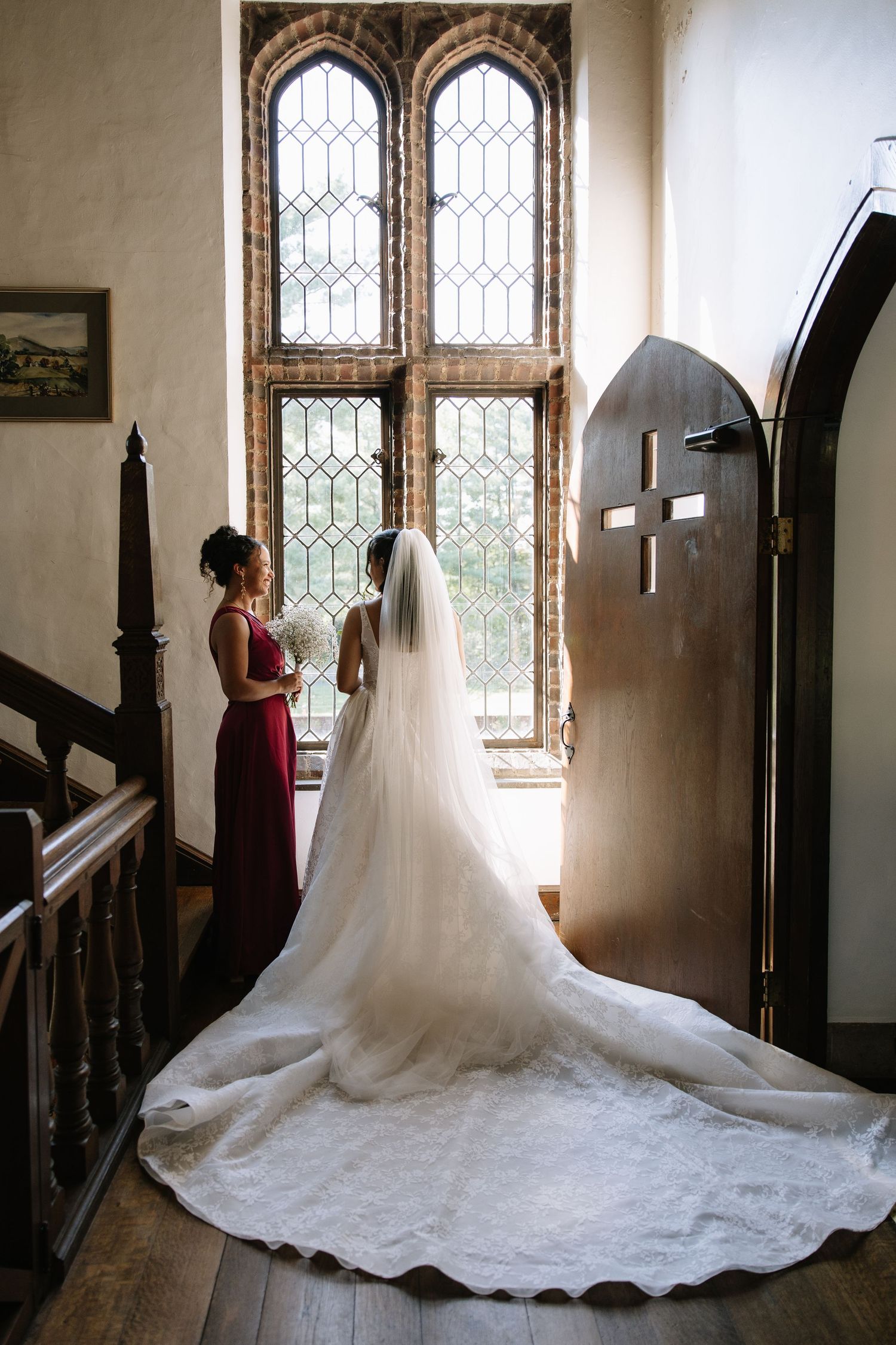 Bride in white gown with long veil stands by gothic leaded windows with bridesmaid in red dress.