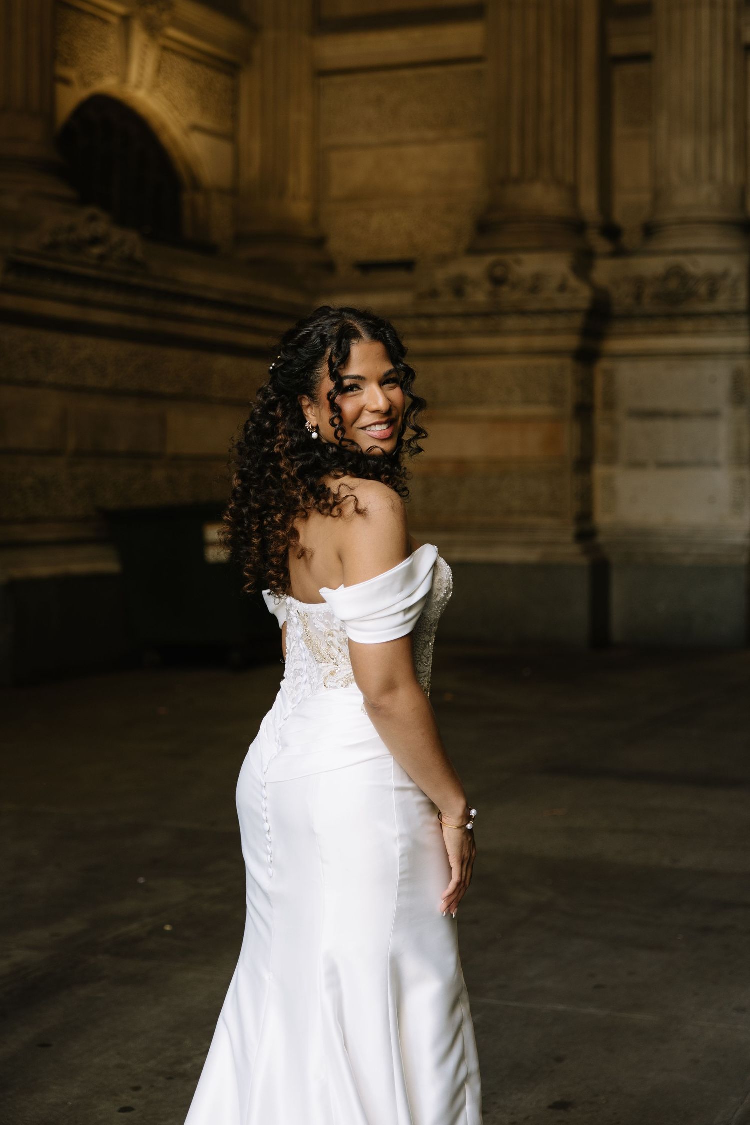 Smiling bride in off-shoulder white lace mermaid gown poses elegantly in front of grand stone architecture.