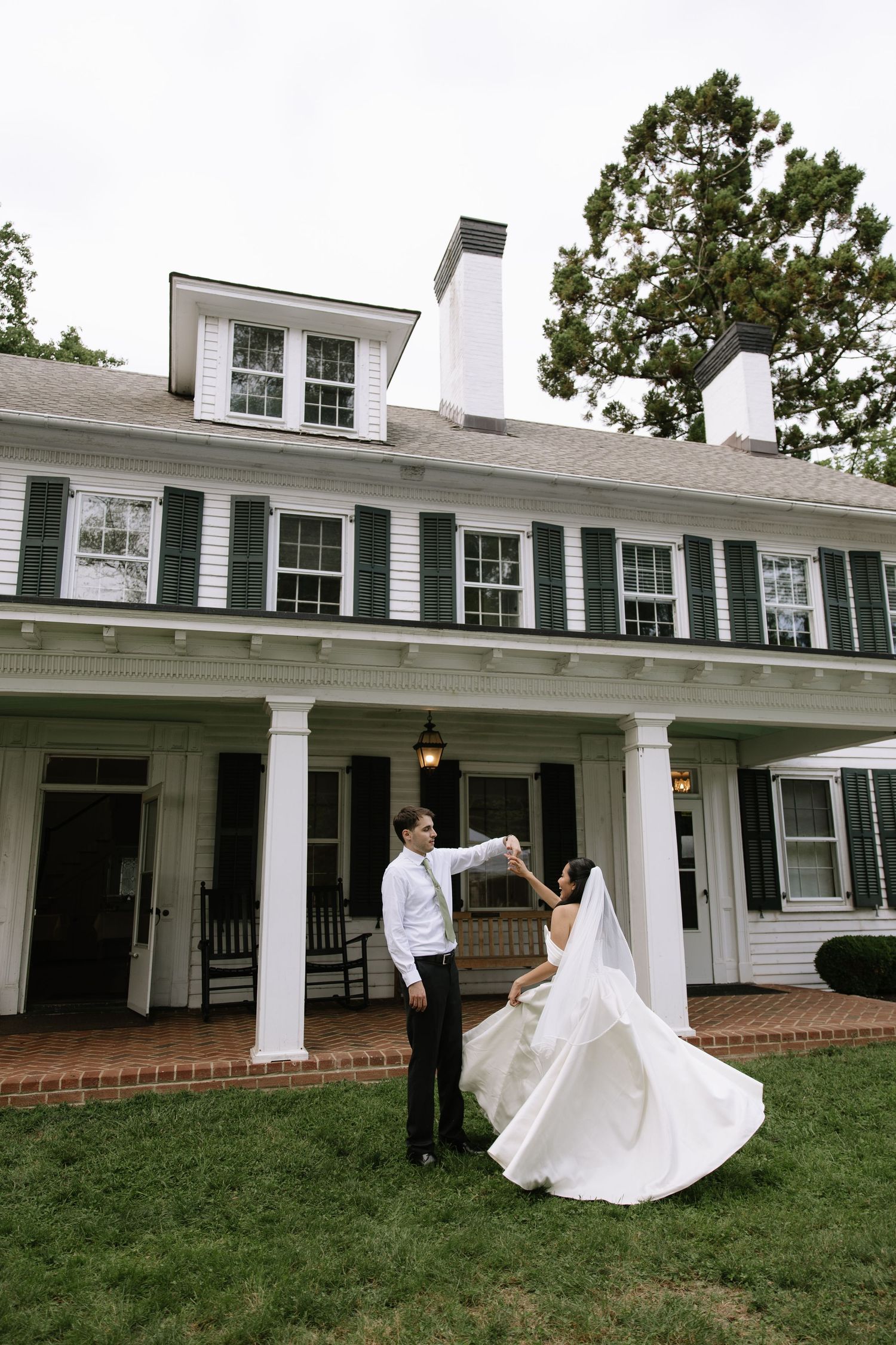 Bride and groom dancing on lawn in front of elegant white colonial home with green shutters and pillars.