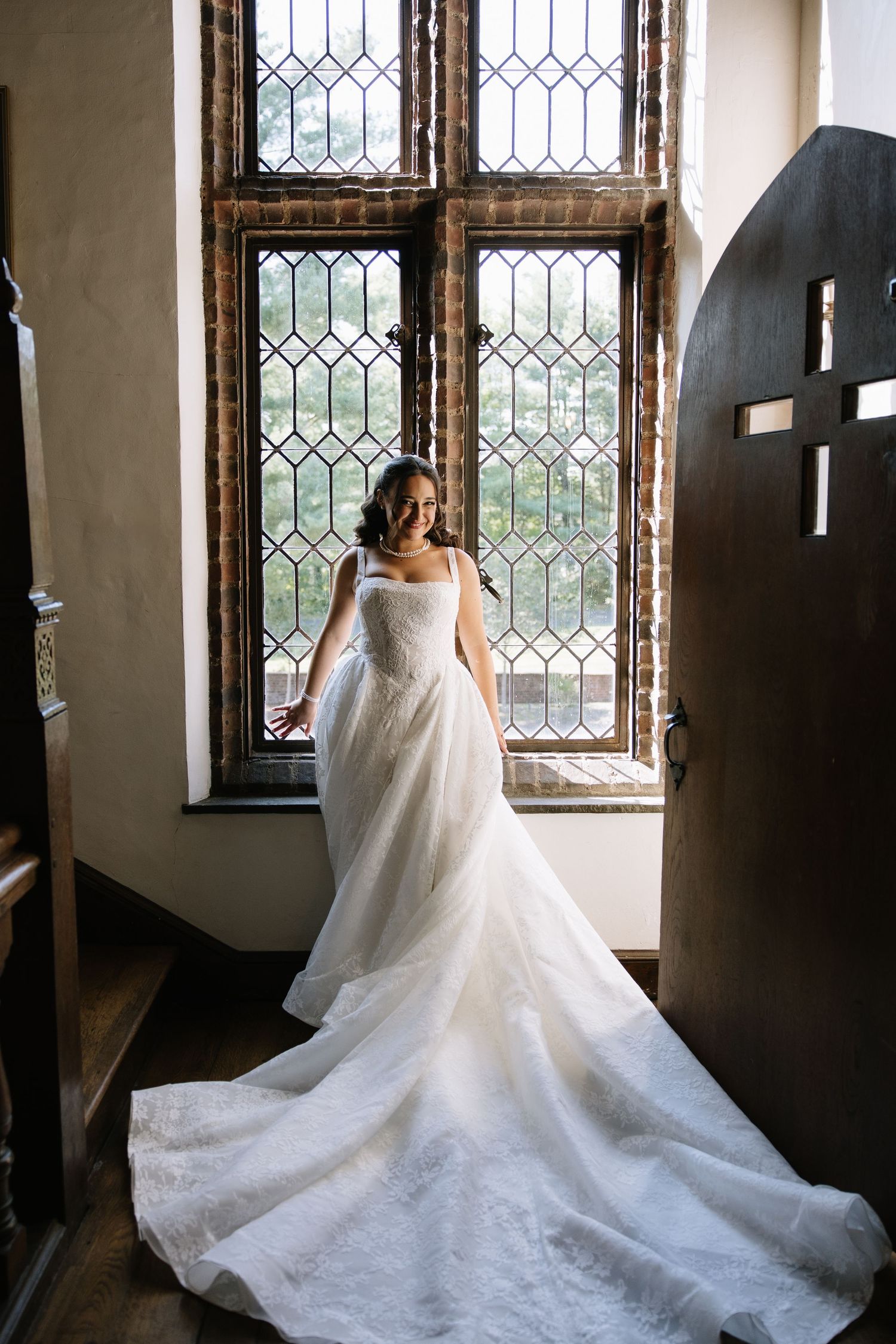 Bride in elegant white gown with long train stands before leaded glass windows in a historic venue.