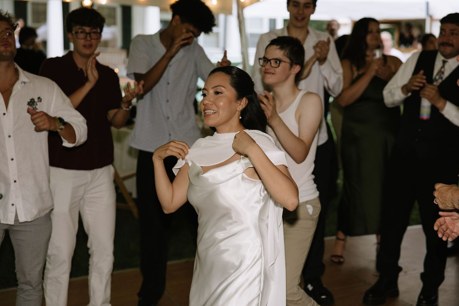 Bride in white satin gown dancing joyfully with guests at wedding reception celebration.