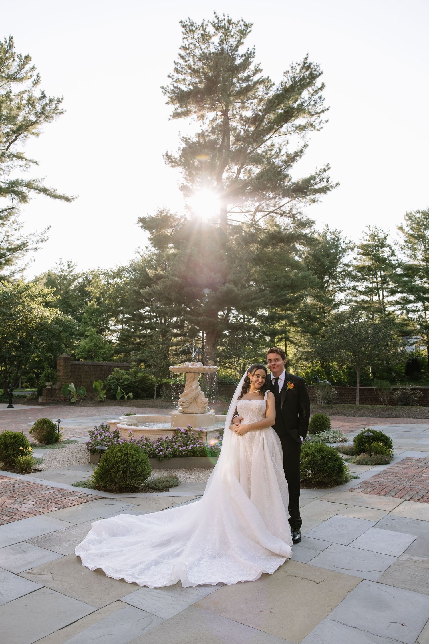 Bride in white ball gown and groom in black tux pose in sunlit garden with stone fountain backdrop.
