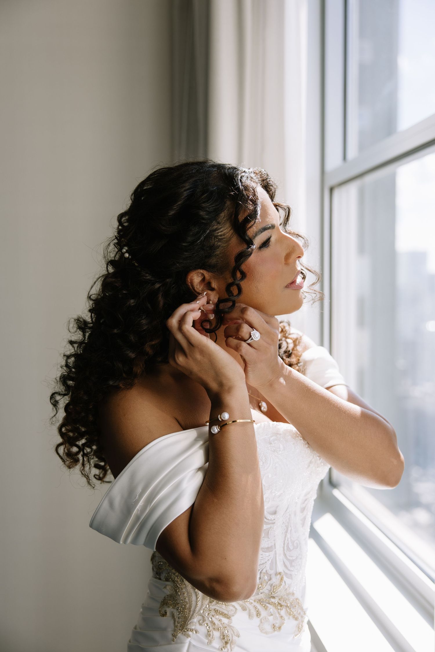 Bride in off-shoulder lace gown putting on earrings by window, curly dark hair adorned with pearl accessories.