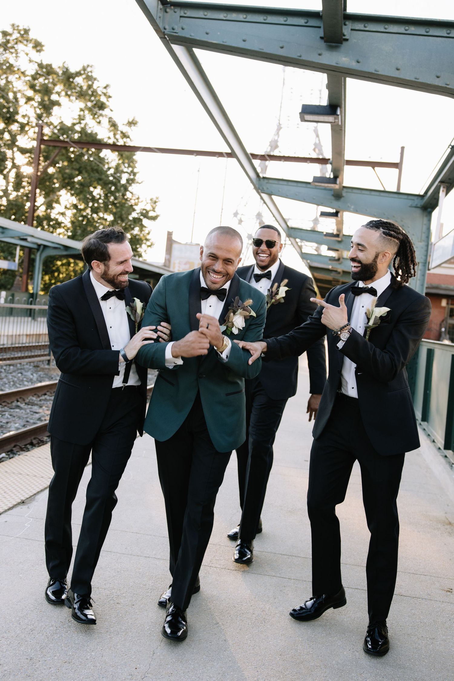 Groom in green tuxedo laughing with groomsmen in black tuxedos on outdoor platform.