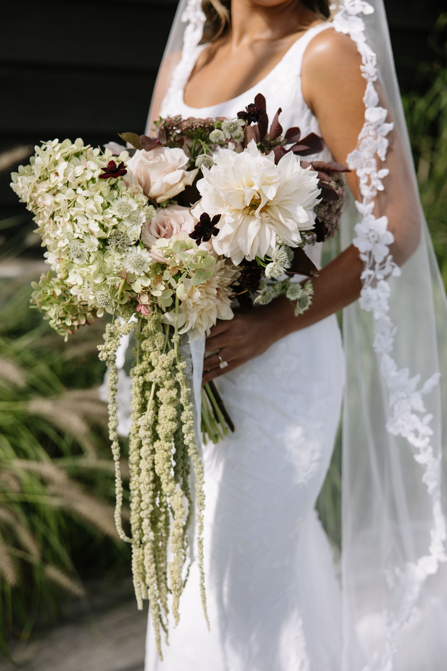 Bride in white gown holding a cascading bouquet of dahlias, roses, and greenery with lace veil.