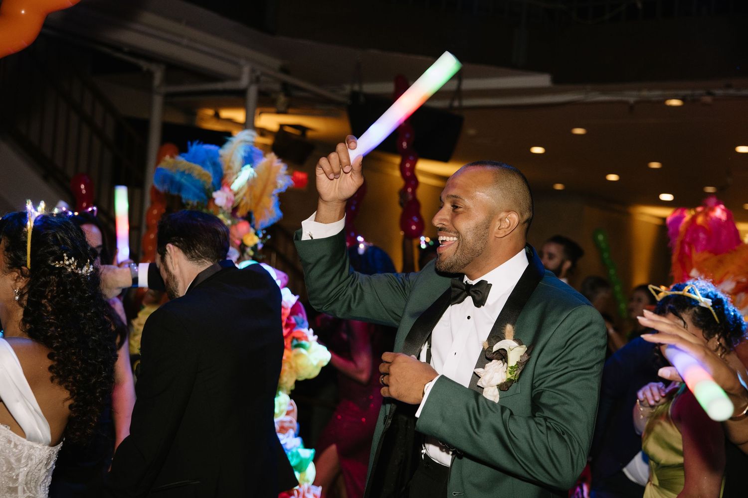 A groom in a teal tuxedo dances joyfully at a wedding reception, holding a glowing light stick.