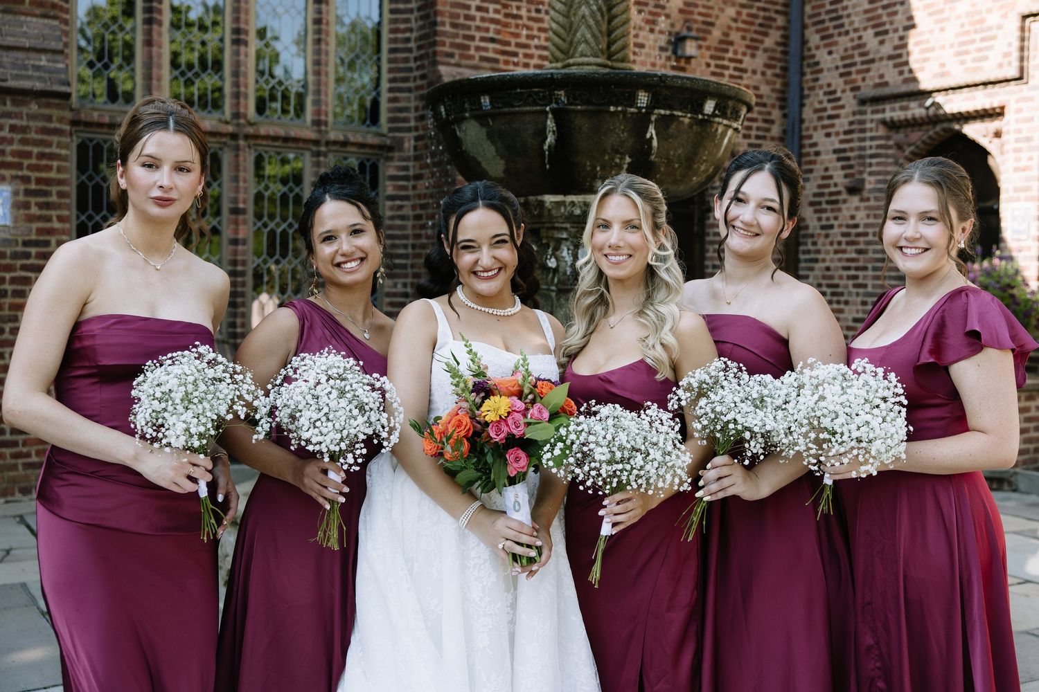 Bride in white gown with five bridesmaids in burgundy dresses holding bouquets, posing outdoors near a brick building.