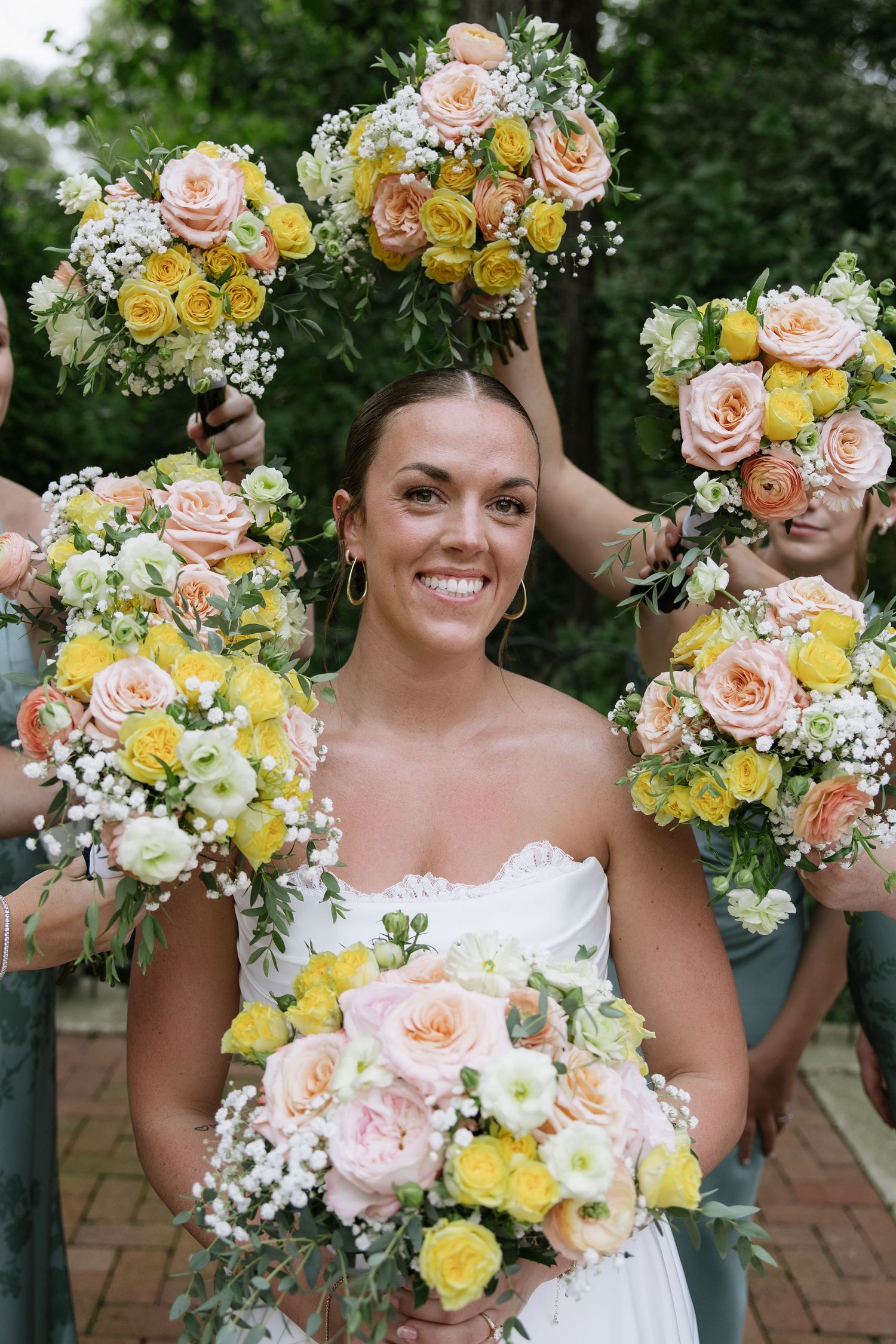 Smiling bride in white dress holds pastel bouquet surrounded by bridesmaids with yellow and pink rose bouquets outdoors.