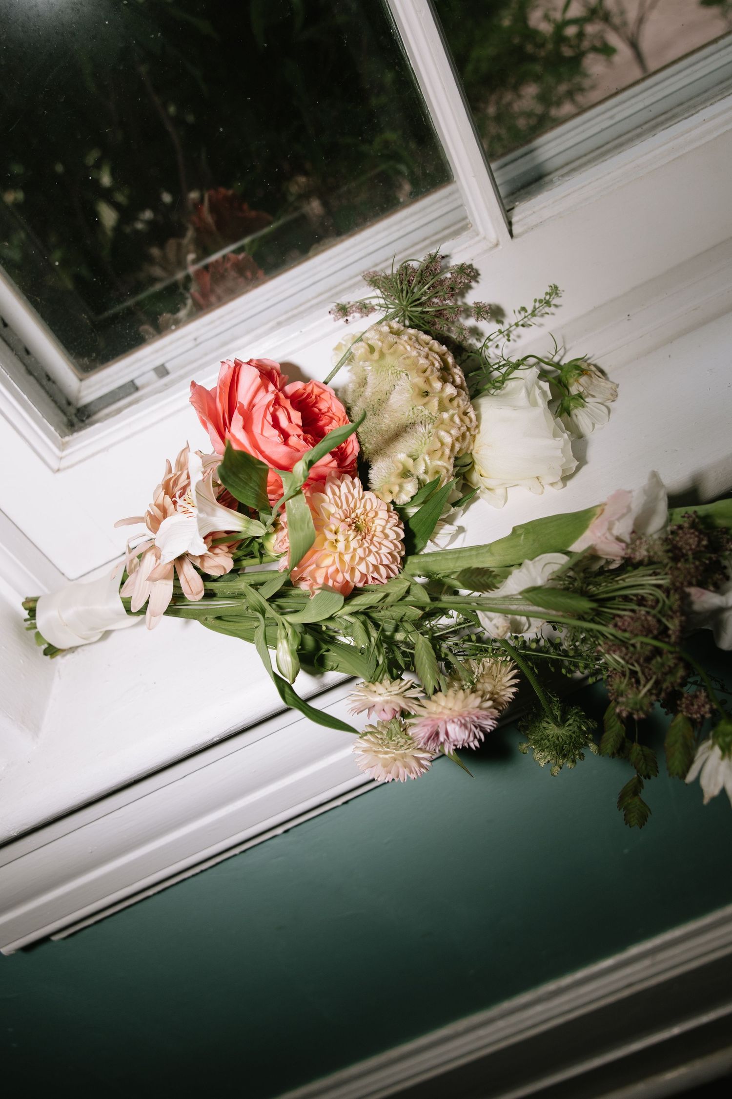 A lush floral bouquet with coral roses, dahlias, and greenery resting on a dark windowsill in soft natural light.
